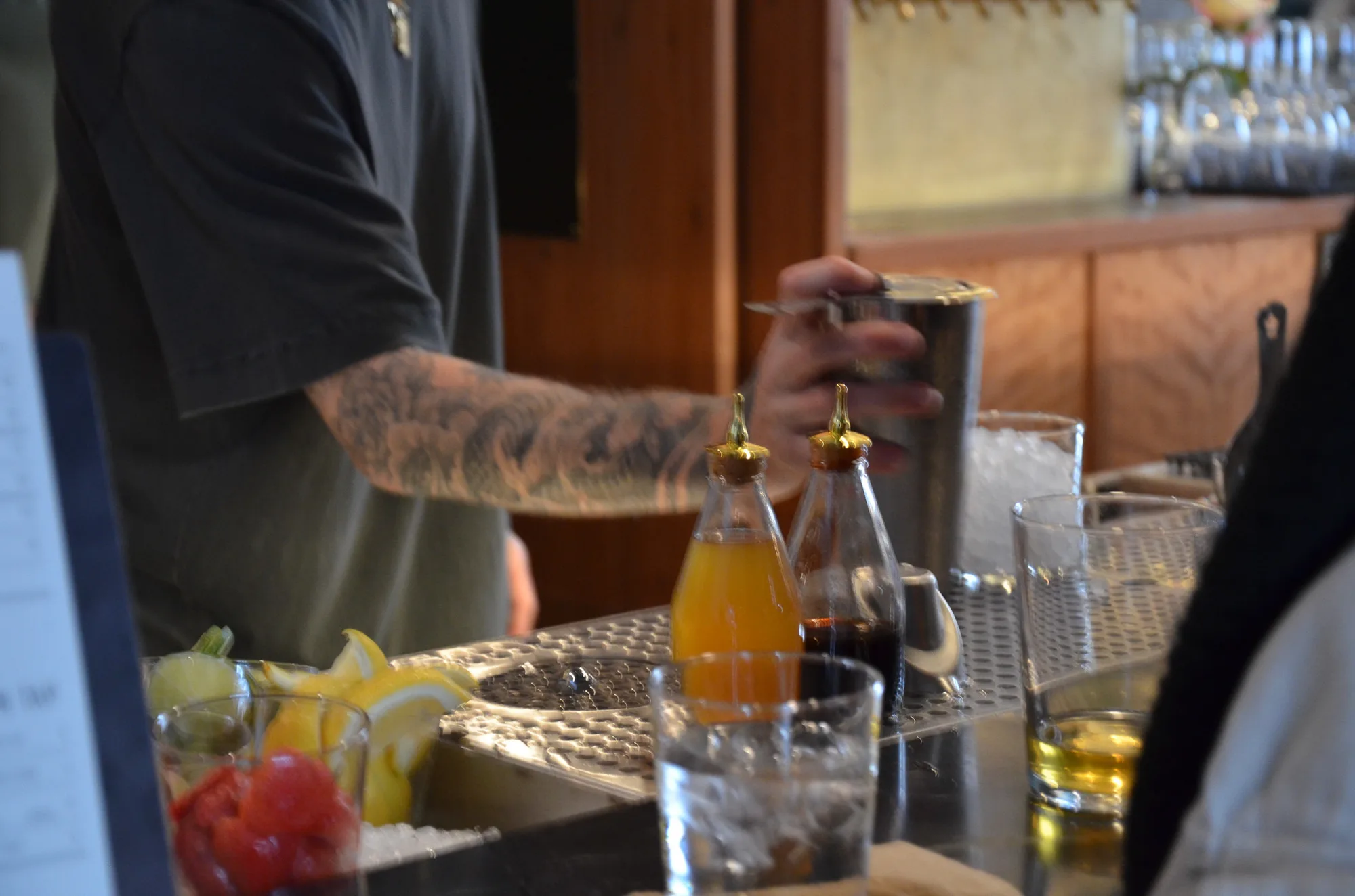 A tattooed bartender pouring at a bar with juices, fruit, and glasses.