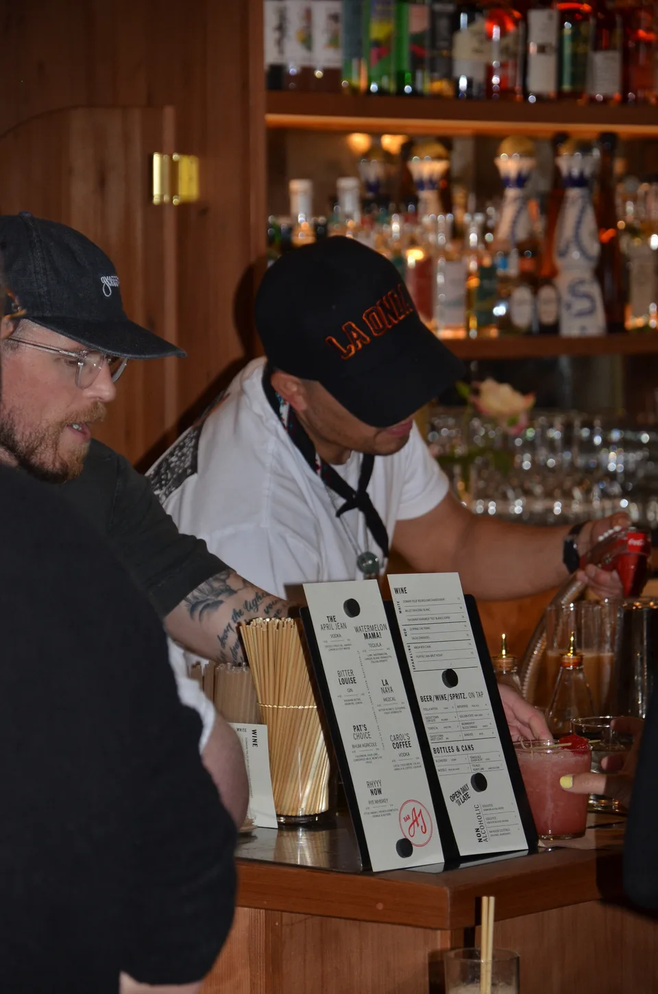 Two bartenders in caps at a bar with printed menus on small stands and a full back bar behind.