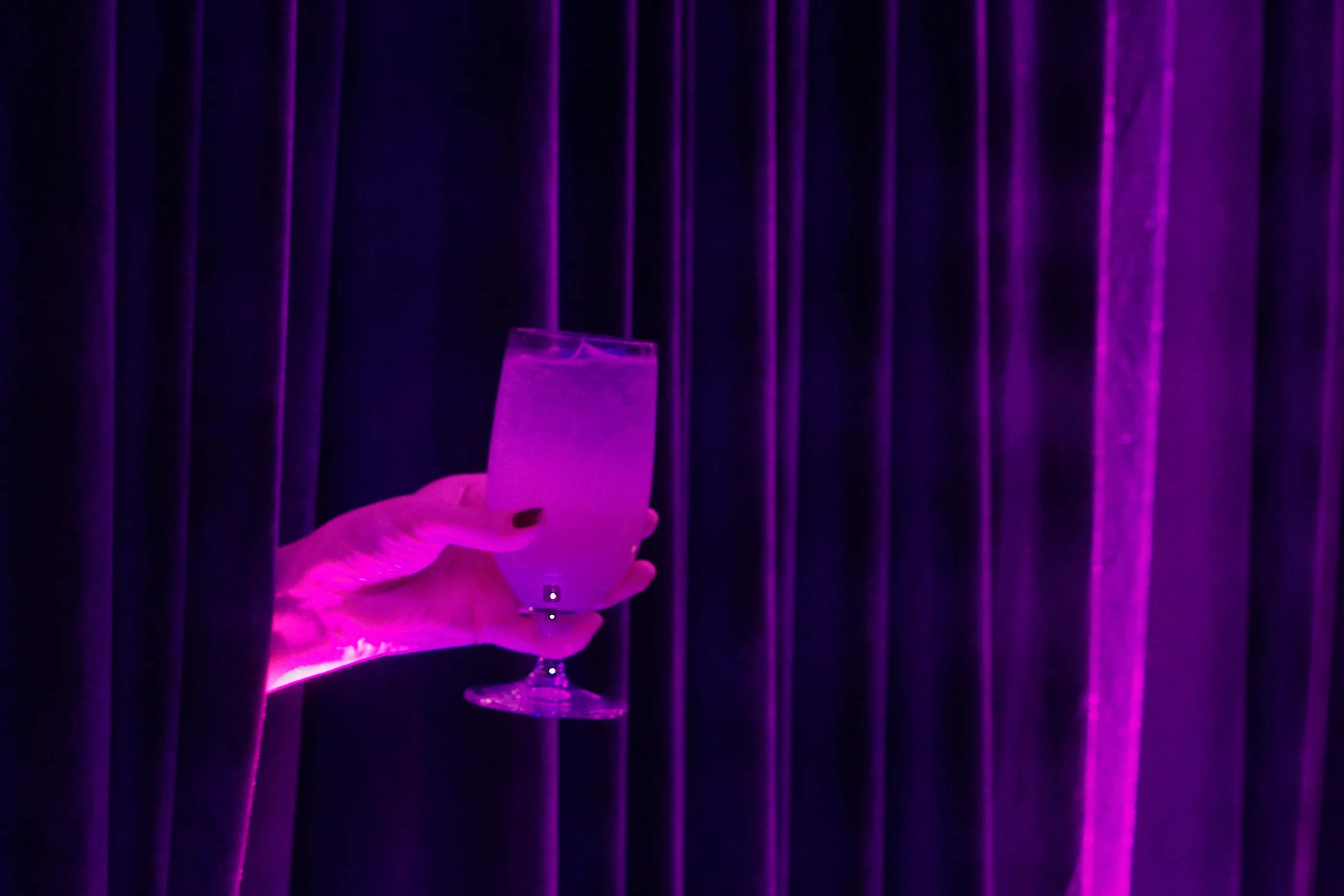 A hand in magenta light holding a glass in front of dark purple velvet curtains.