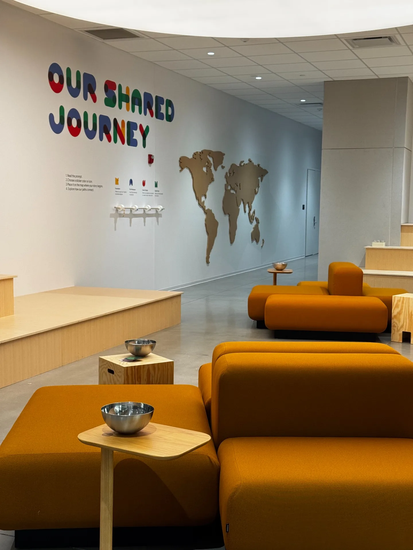 Colorful Our Shared Journey lettering and a world map above timber benches and orange seating.
