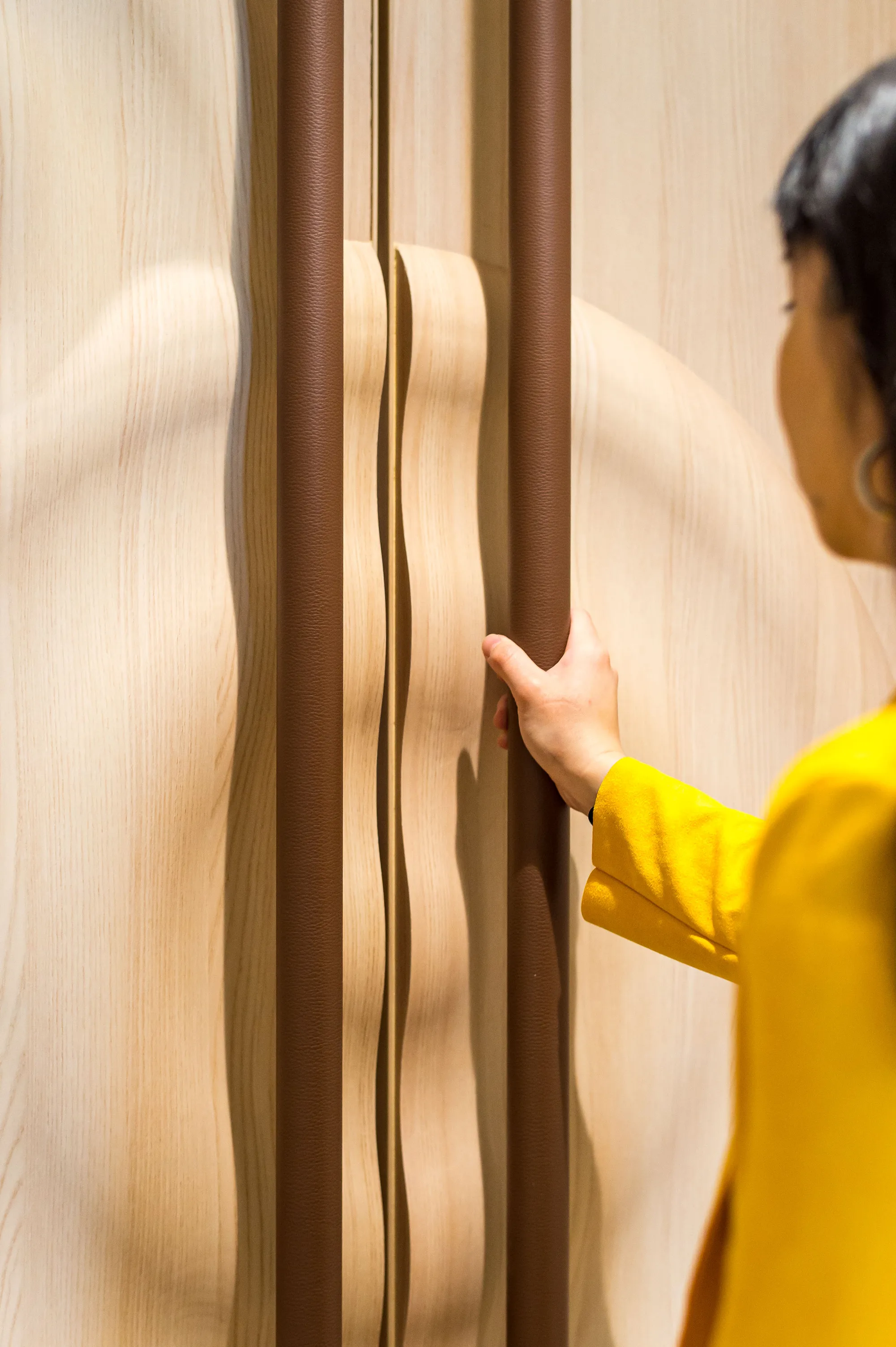 A hand in a yellow sleeve pulling a tall wooden door with a leather-wrapped handle.