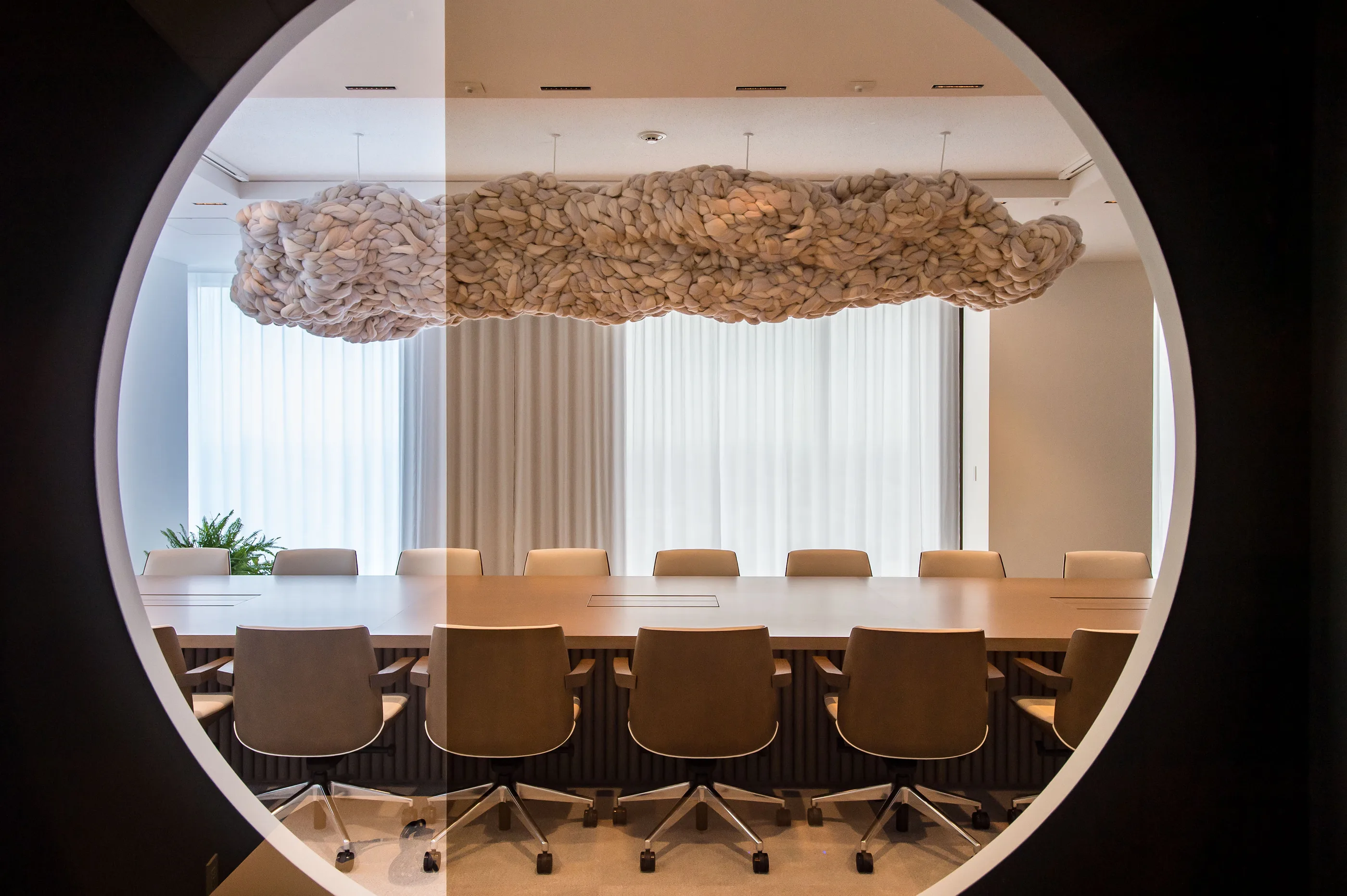 View through a circular window into a meeting room with a long table and a textured white sculpture above.