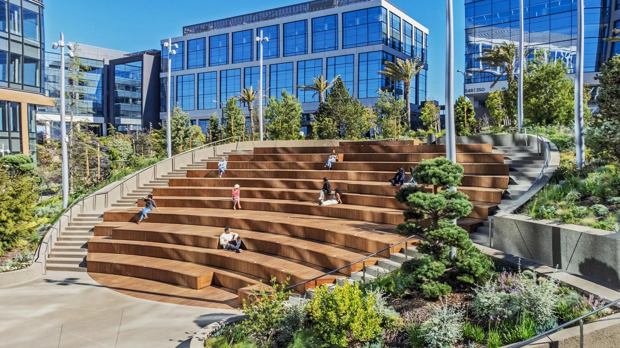 Timber amphitheater steps with people seated, a stair beside them and office buildings behind.