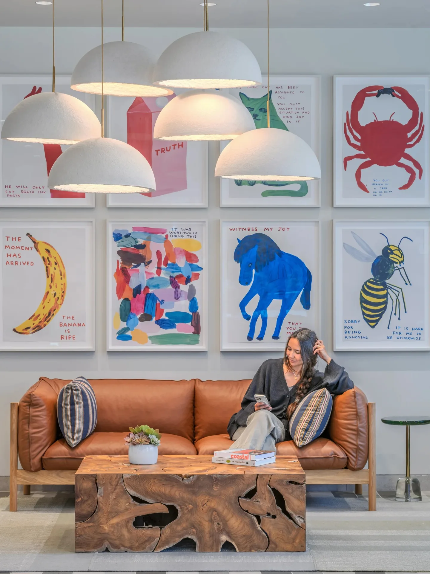 A person seated on a leather sofa with a root-wood table, under white dome pendants and a wall of framed animal prints.