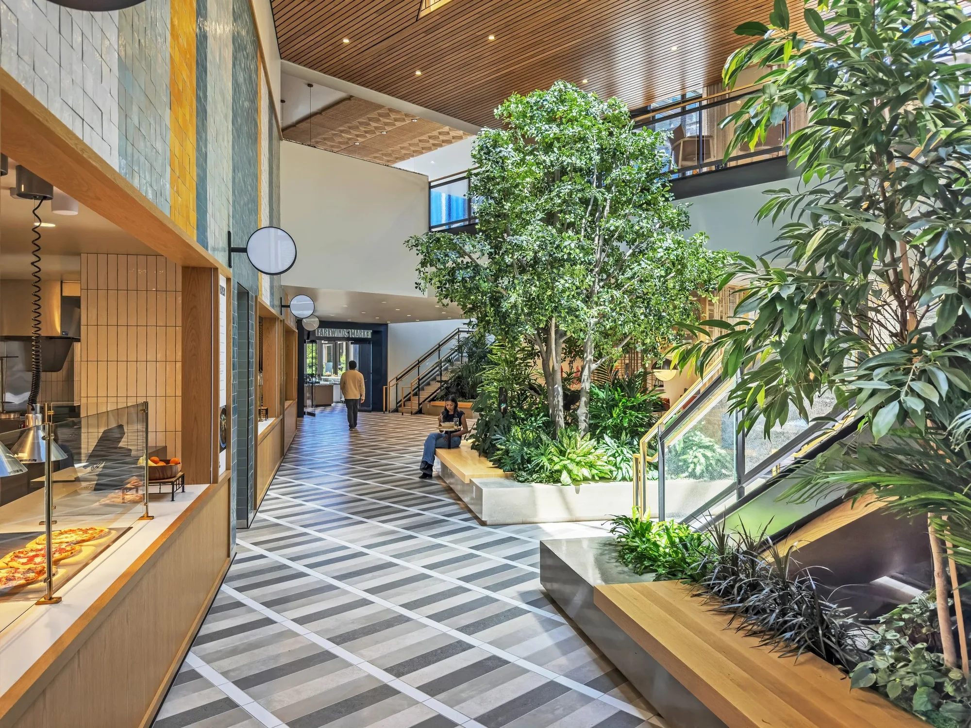 Double-height food hall atrium with a tree in a planted bed, a patterned floor, timber ceiling and a stair.