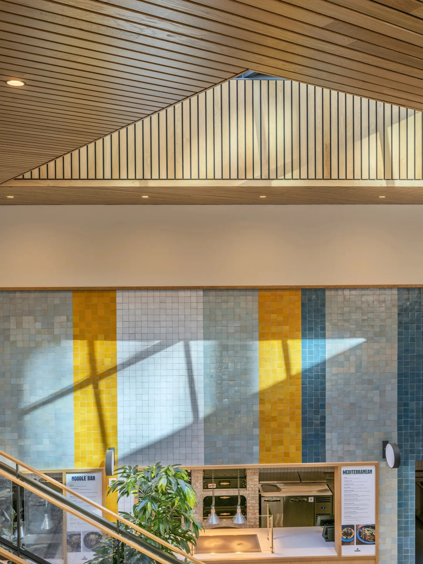 Tiled wall with yellow and blue vertical stripes beneath a pitched timber ceiling, with a stair rail below.