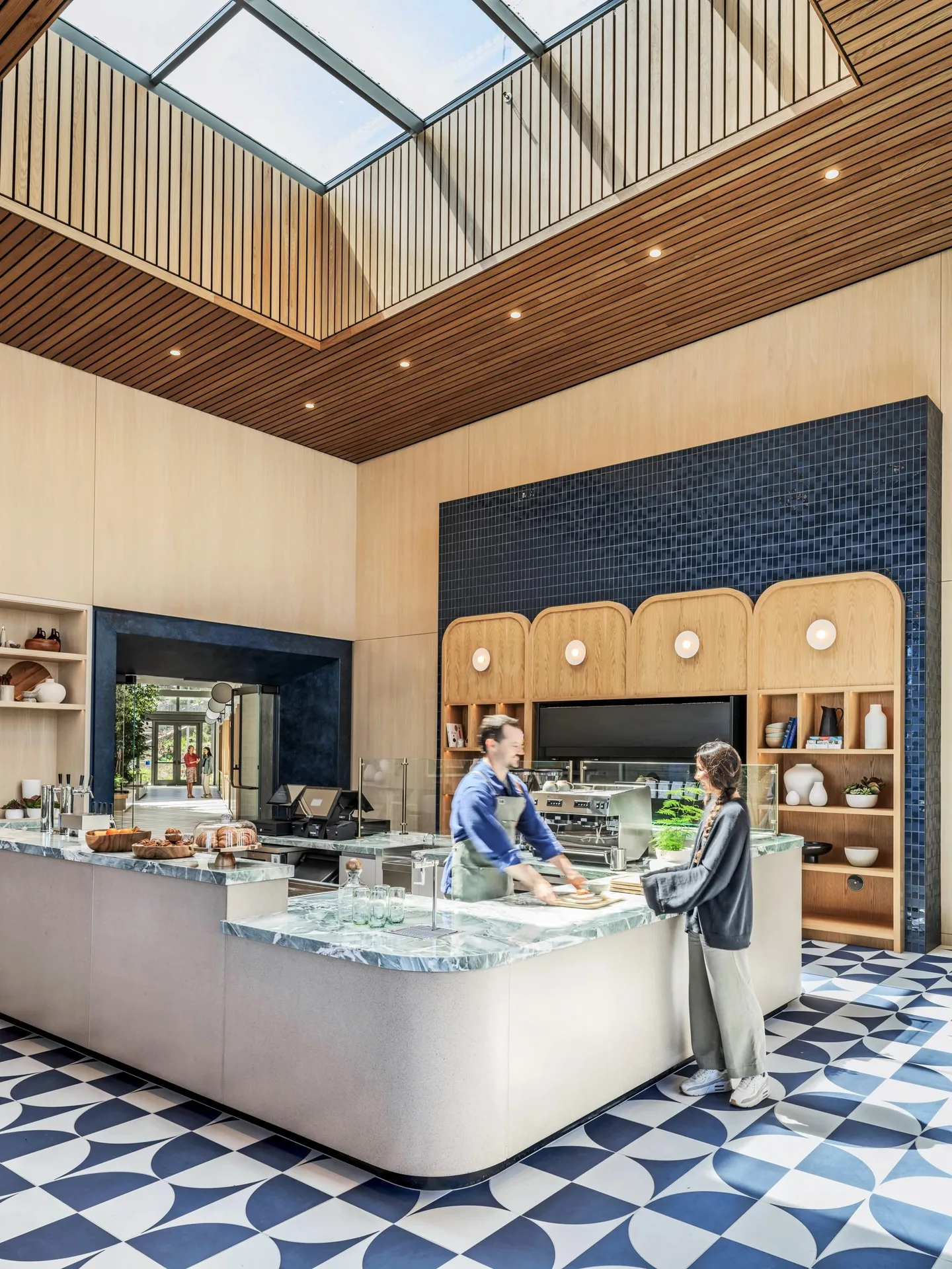 A cafe counter in pale stone with blue and white geometric tile, arched shelving and a skylight above.