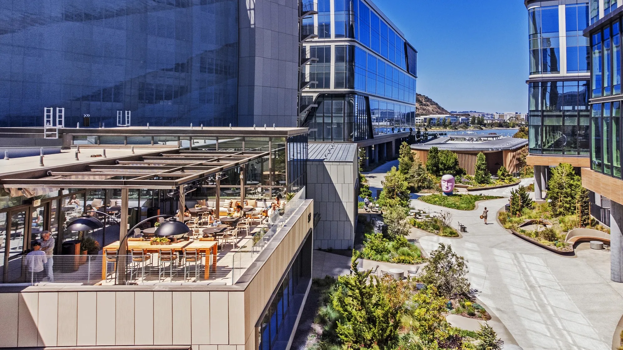 Aerial view of a terrace with dining tables beside the building, with campus paths, planting and glass buildings beyond.