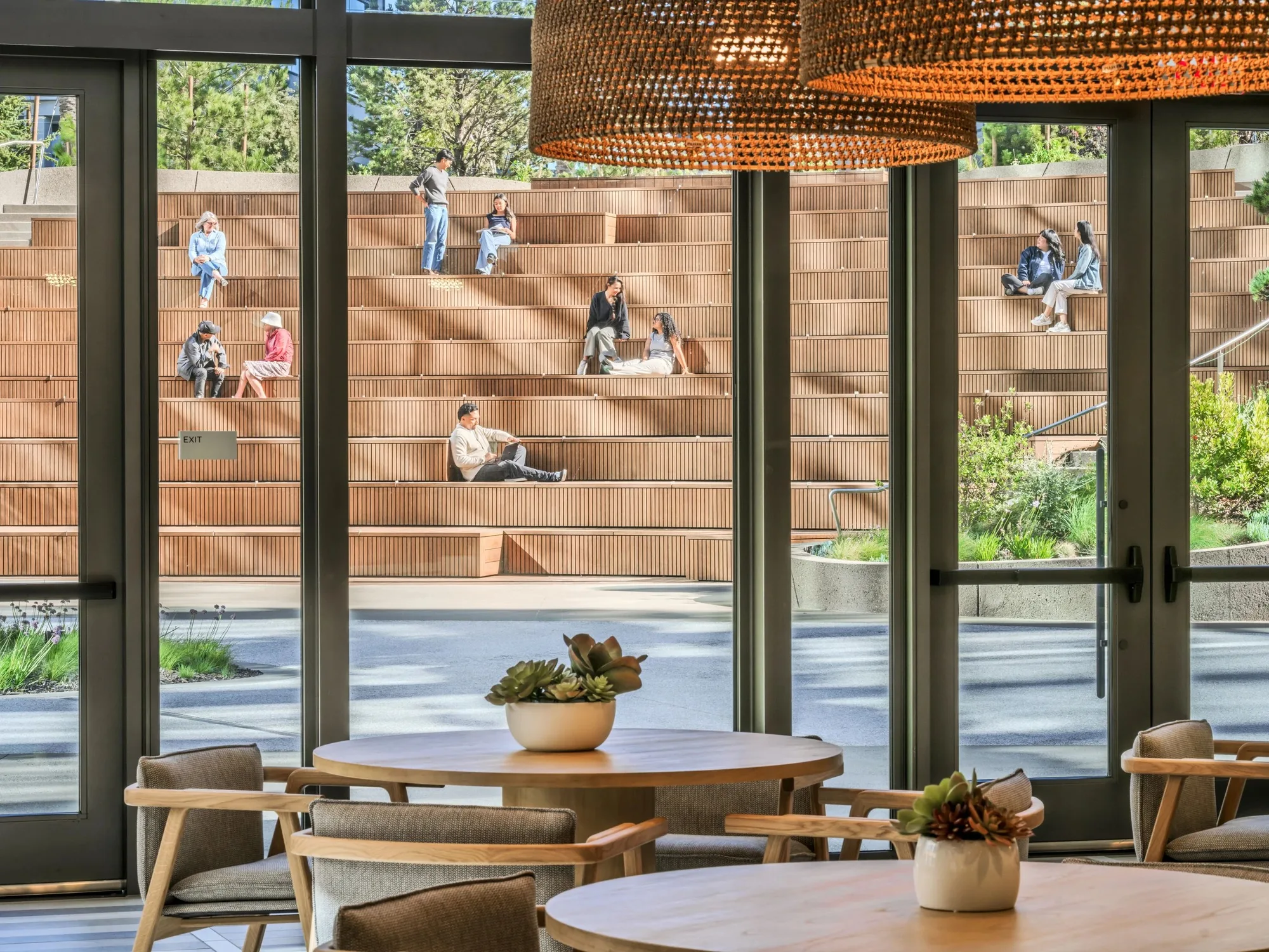 Dining tables with woven pendant lights behind tall glass doors, with the timber amphitheater and people outside.