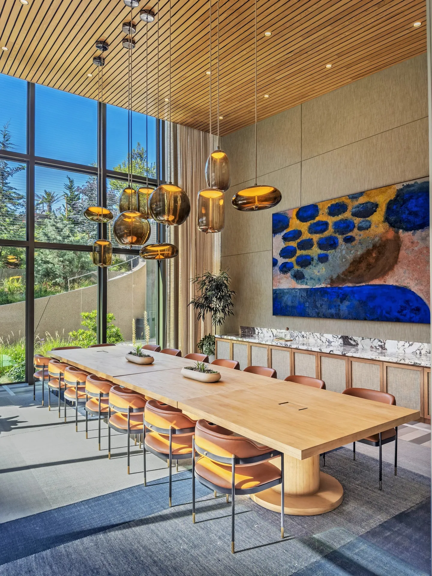 A long timber table with orange chairs under amber pendant lights, with tall windows and a blue and yellow painting.