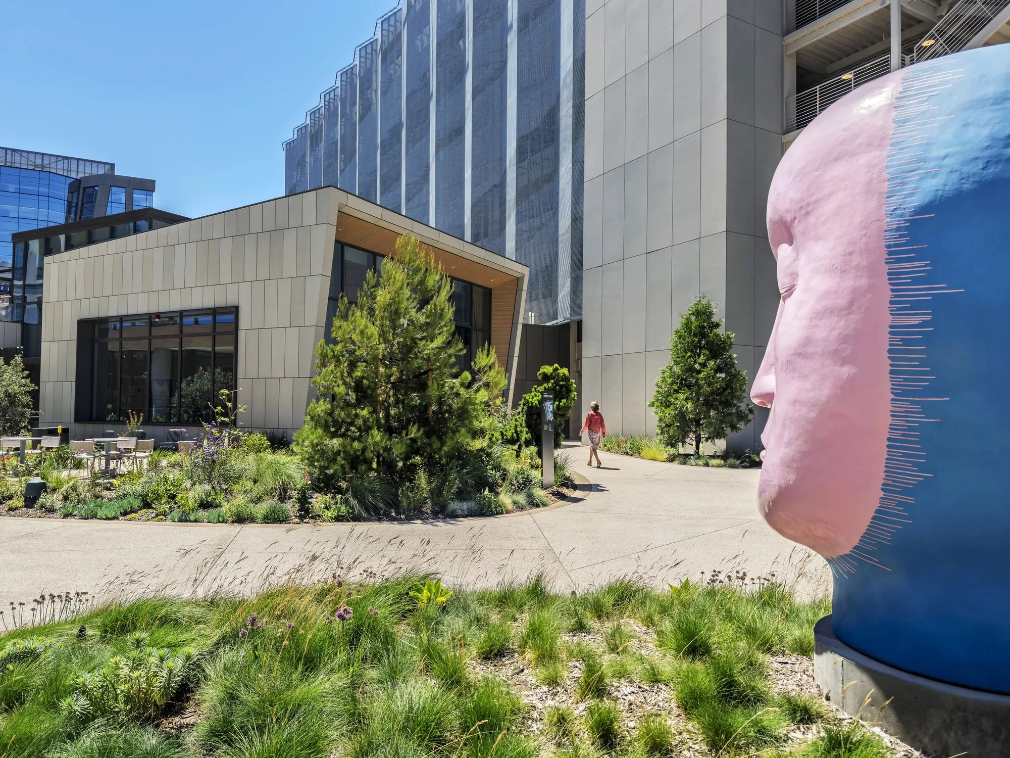 A low building with a sloped roof among planting, with a large pink and blue sculpted head in the foreground.