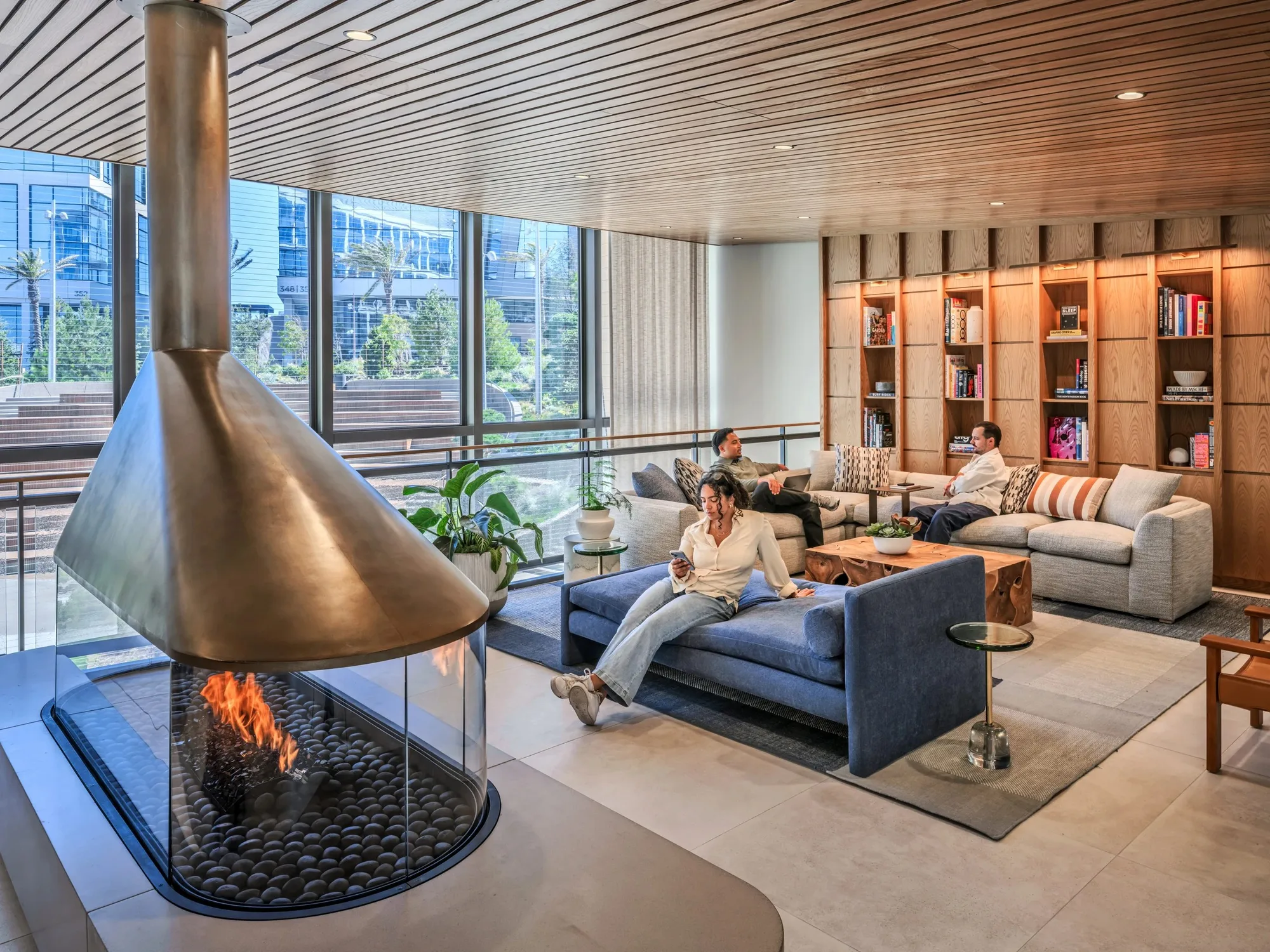 Lounge with a suspended conical brass fireplace, sofas, a timber ceiling and shelving, with people seated.