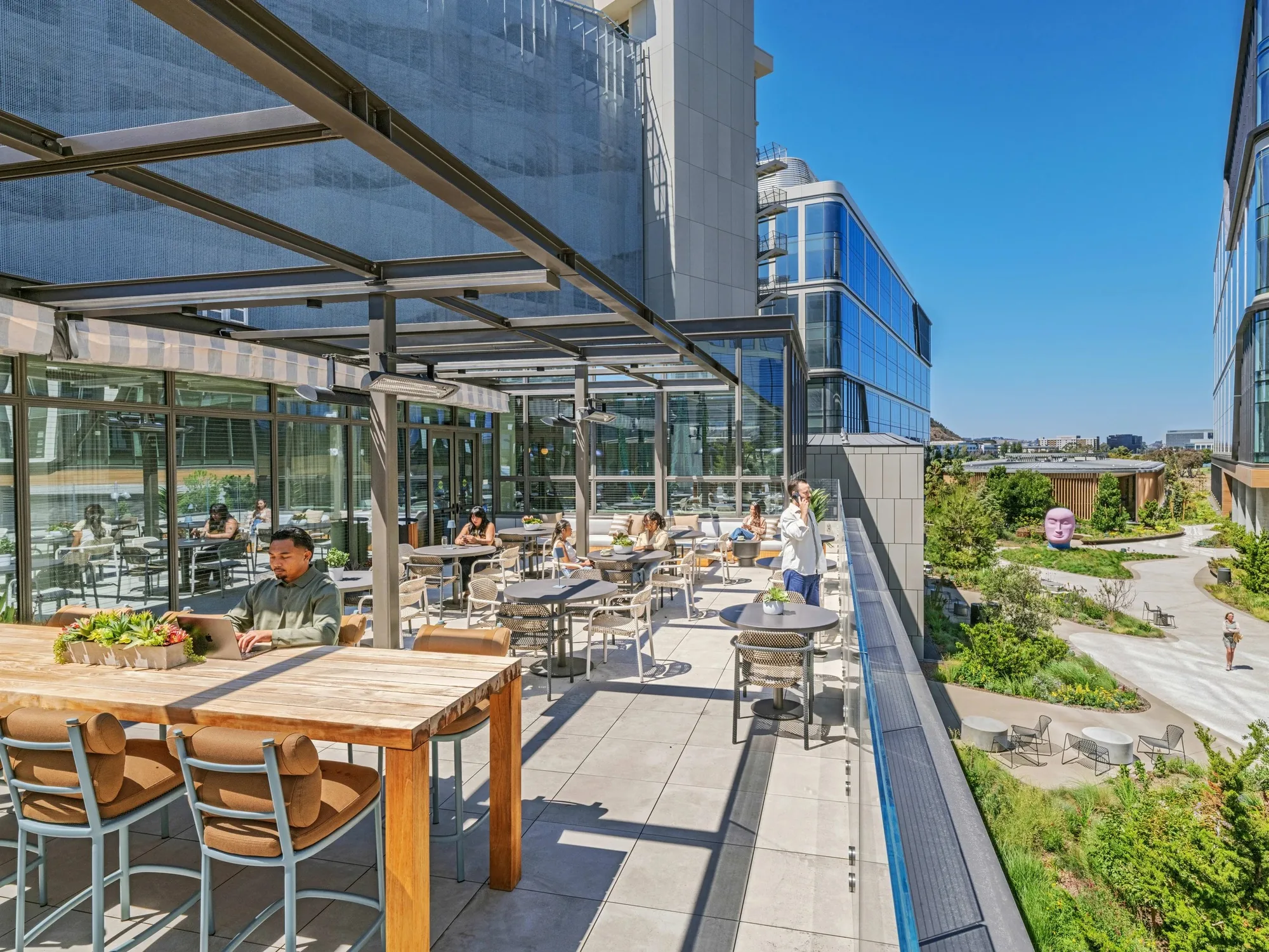 A covered terrace with timber and metal tables and chairs and people dining, overlooking planting and glass buildings.