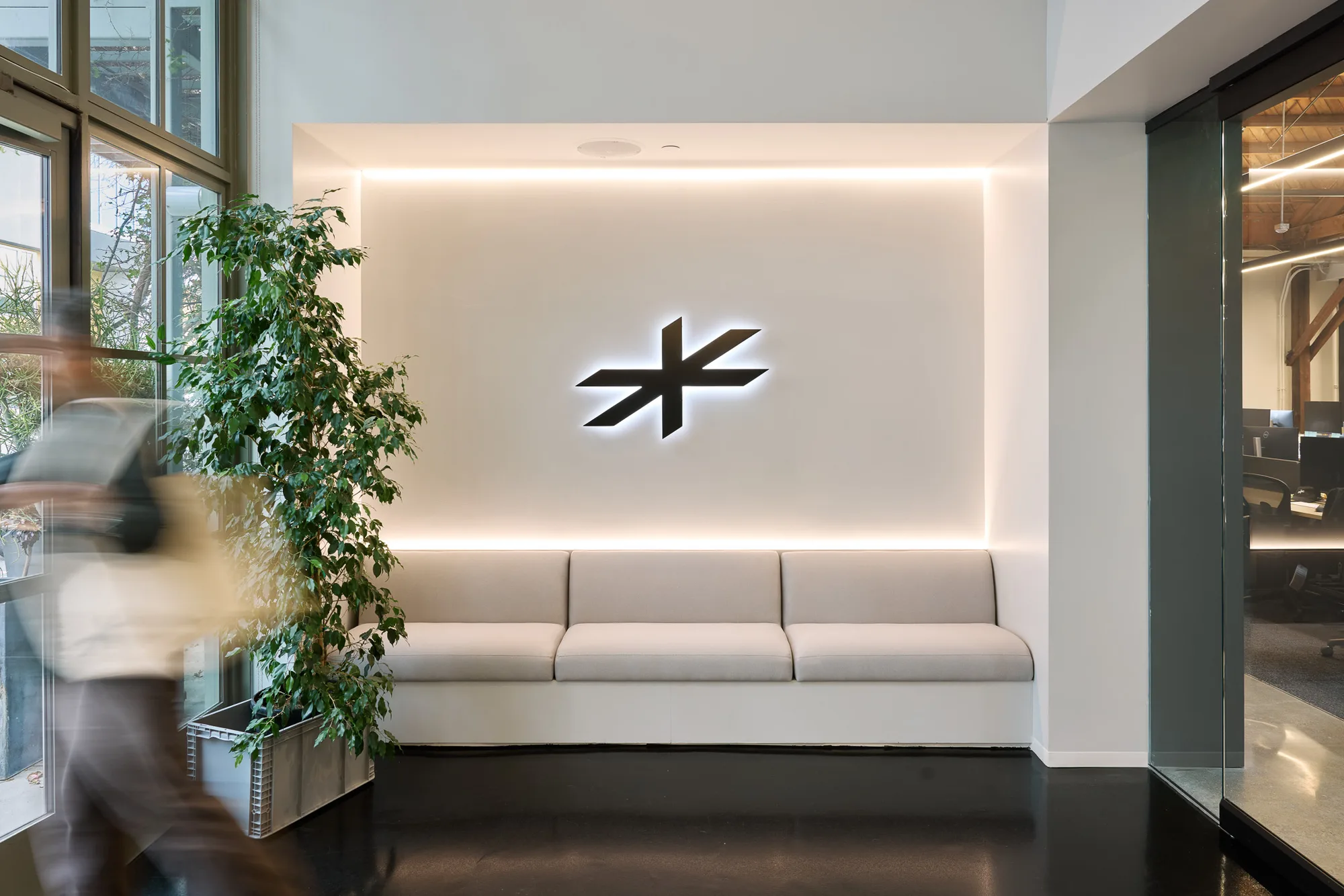 A pale bench seat on a white wall with a backlit asterisk-shaped mark, beside a large plant and a person walking past.