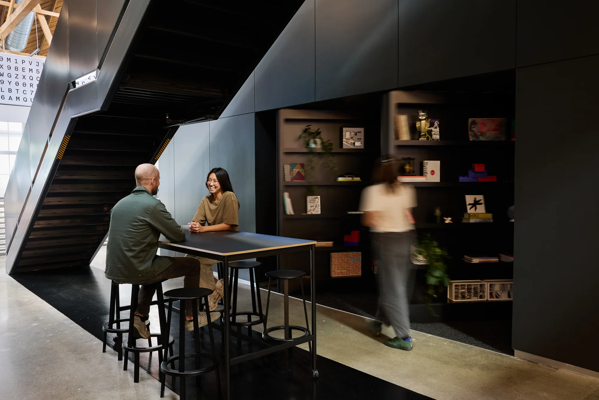 Two people talking at a tall black table beside a black stair and shelves, with a third person passing.