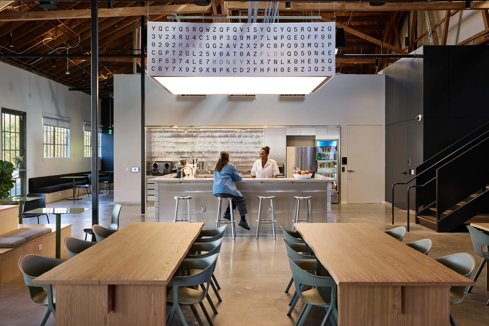A kitchen counter with two people and long timber tables below a white light panel printed with a grid of letters.