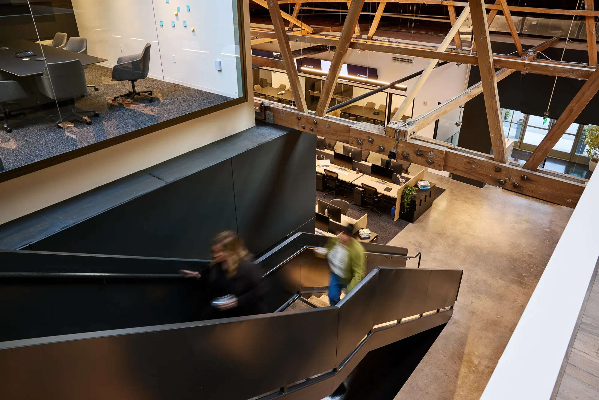 View from above of a black stair, timber trusses, and desks on the floor below.