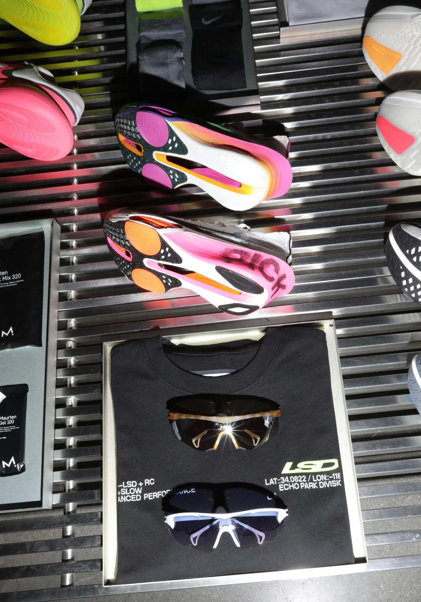 Overhead view of running shoes, a black LSD tee and sunglasses on a slatted metal surface.