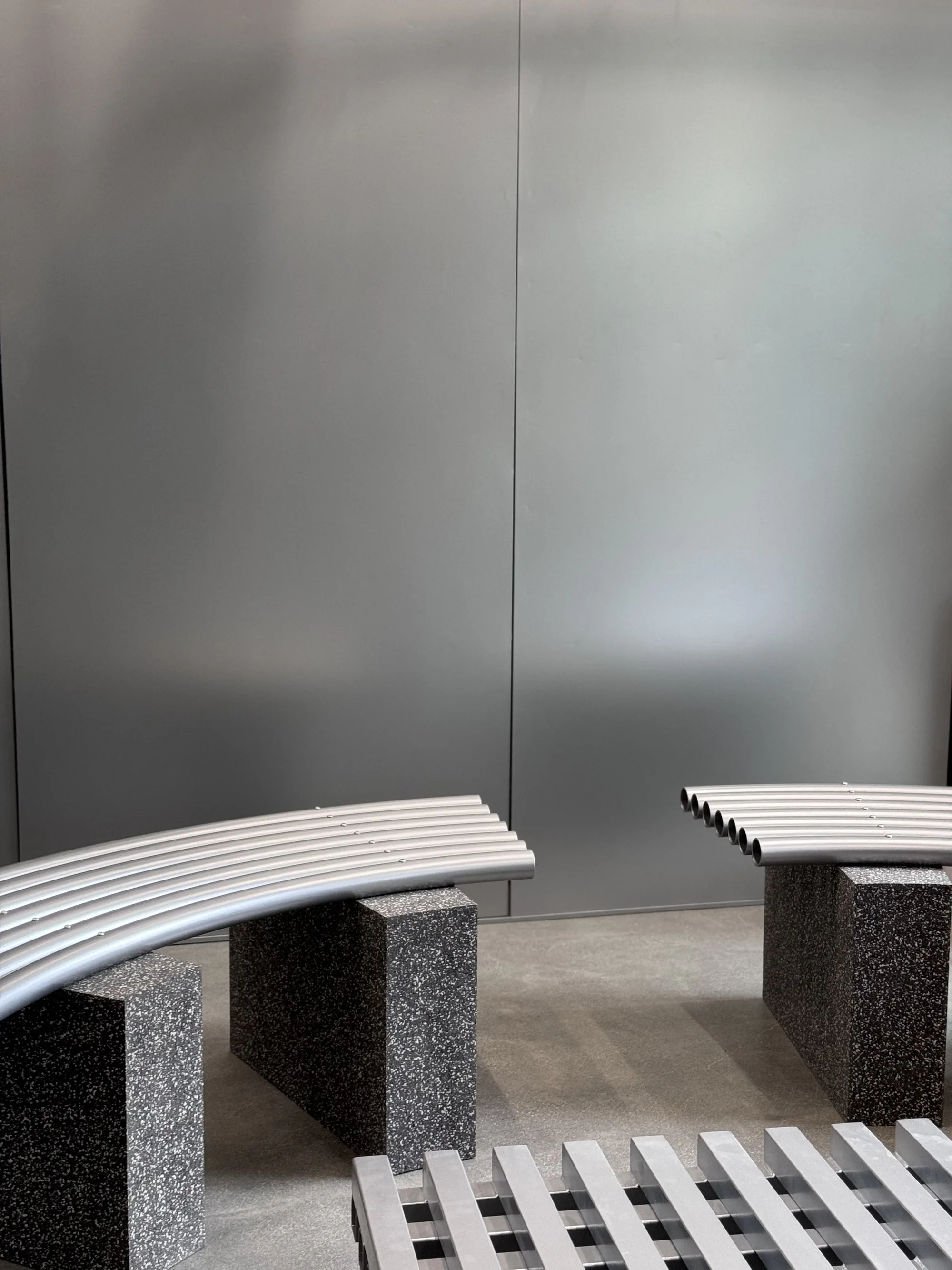 Curved tube bench on stone supports in front of a brushed metal wall.