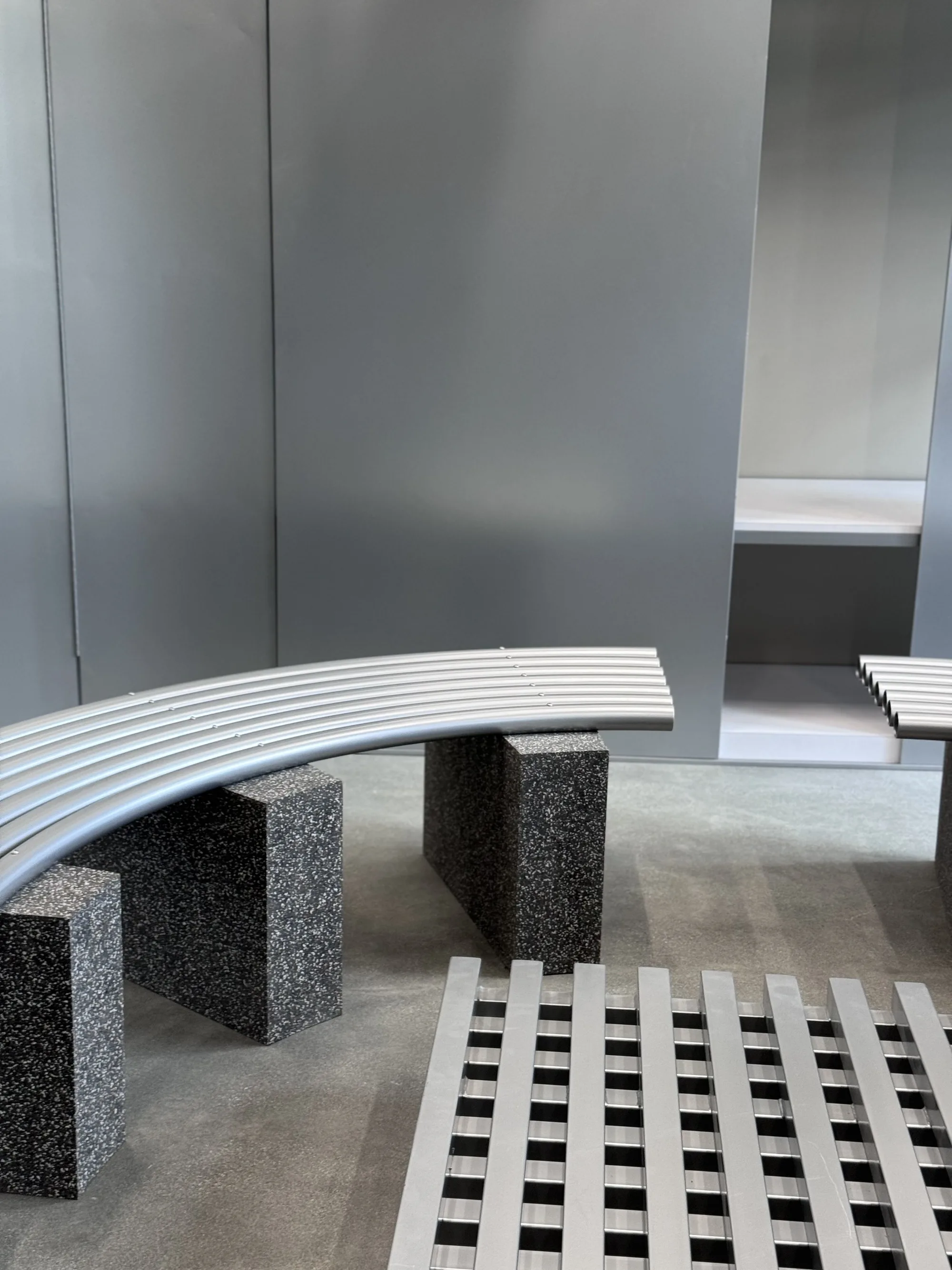 Curved tube bench on stone blocks beside a slatted platform in front of a metal wall.