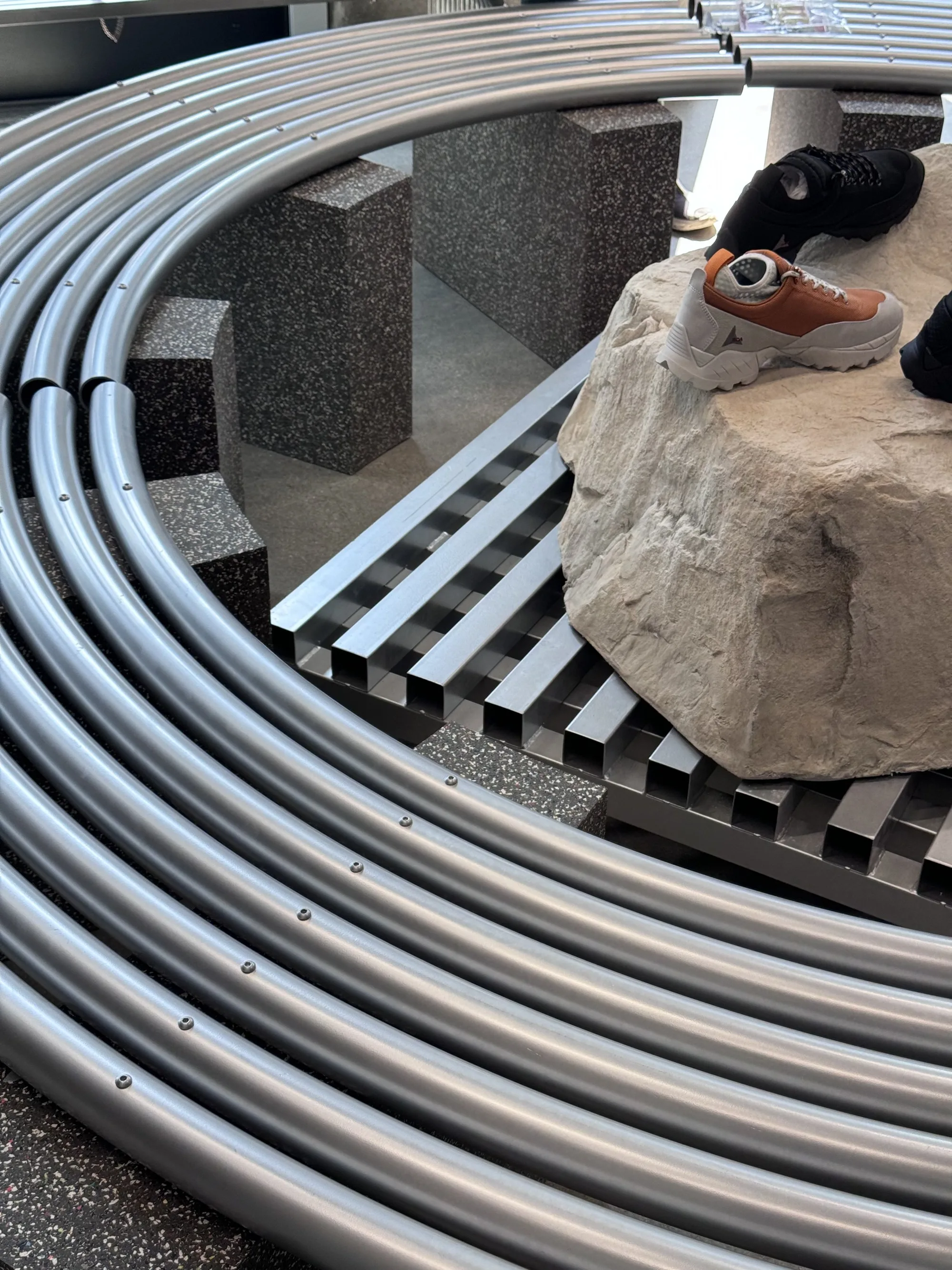 A rock display with a shoe on top, surrounded by concentric curved metal tubes and a slatted platform.