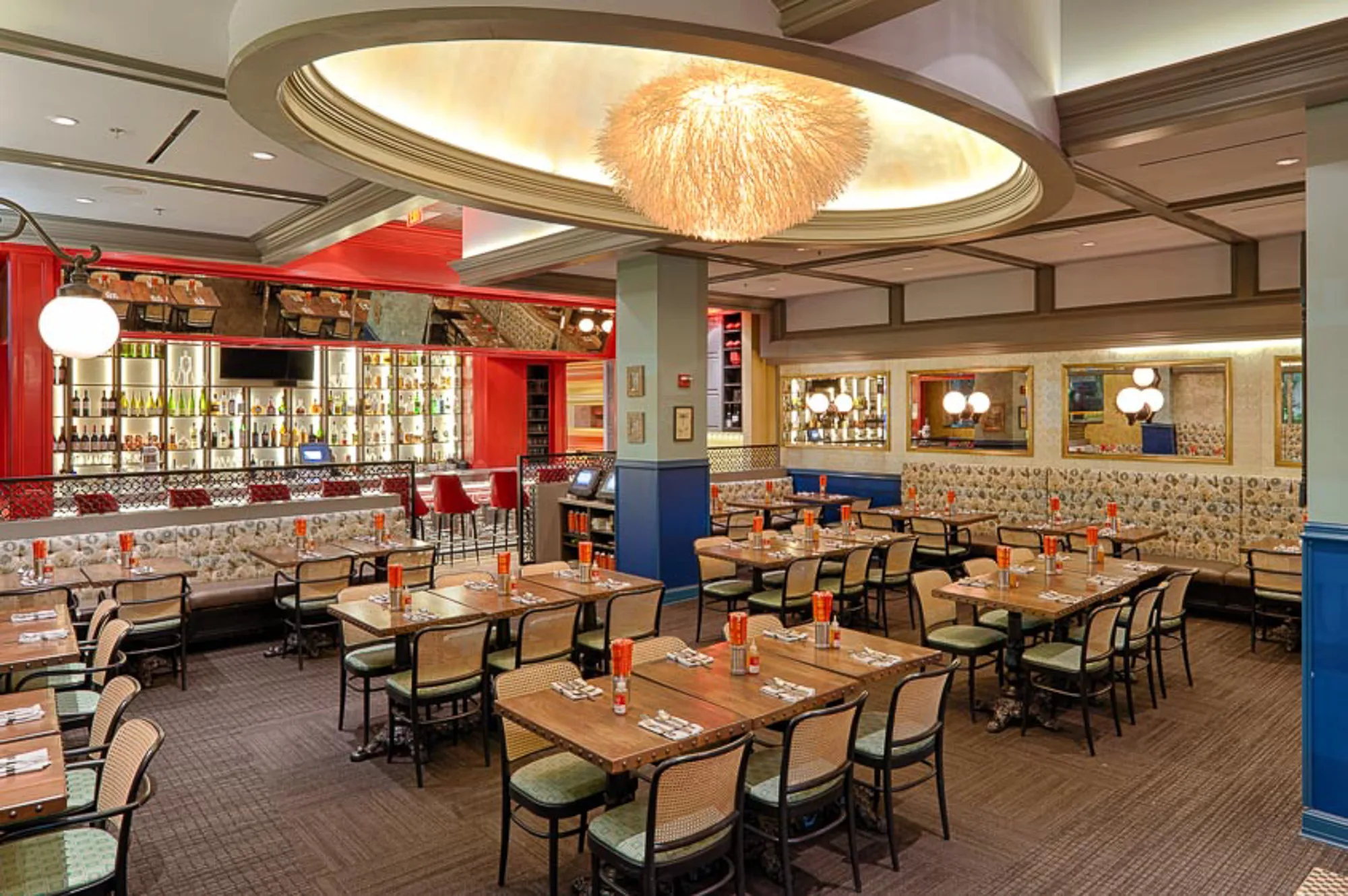 Wide view of a restaurant with a chandelier in an oval ceiling, red bar, and many set tables.