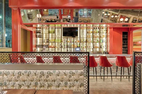 A bar with red stools below shelves of bottles, in front of a patterned banquette.