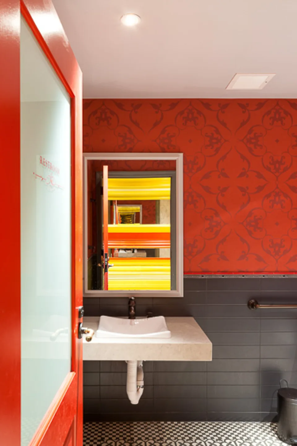 Restroom with red walls, a white basin, and a mirror showing bands of yellow and red light.
