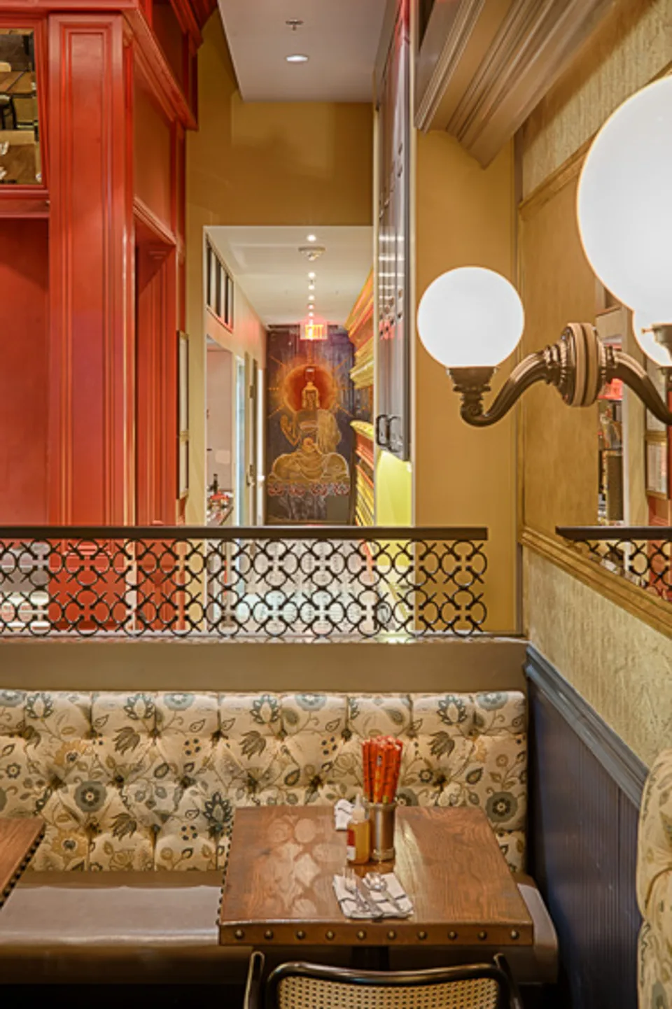 A banquette and table in warm tones with a wall lamp, and a corridor with a painted figure in the distance.