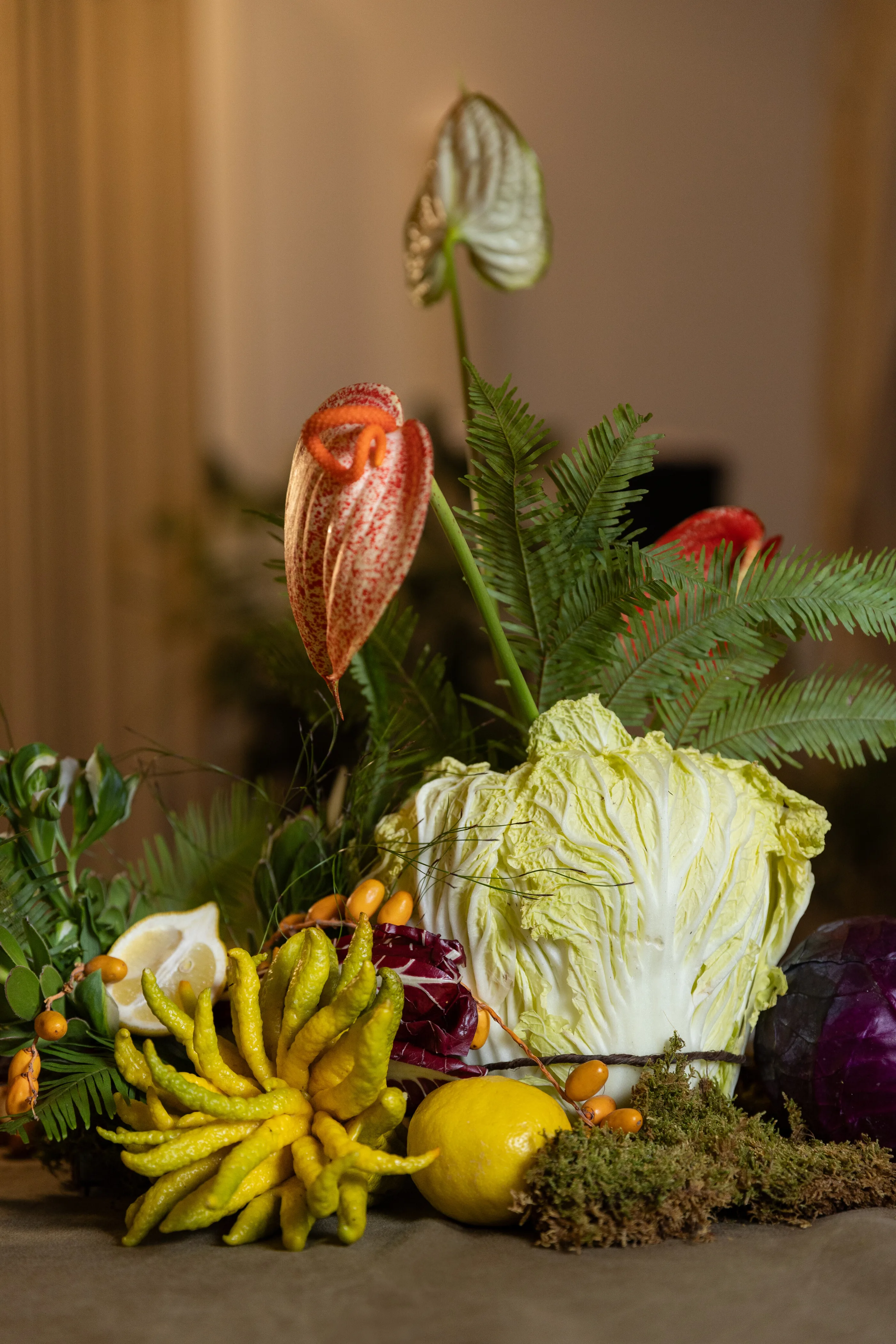 A centerpiece of anthurium, ferns, a pale cabbage, a citron, and moss on a table.