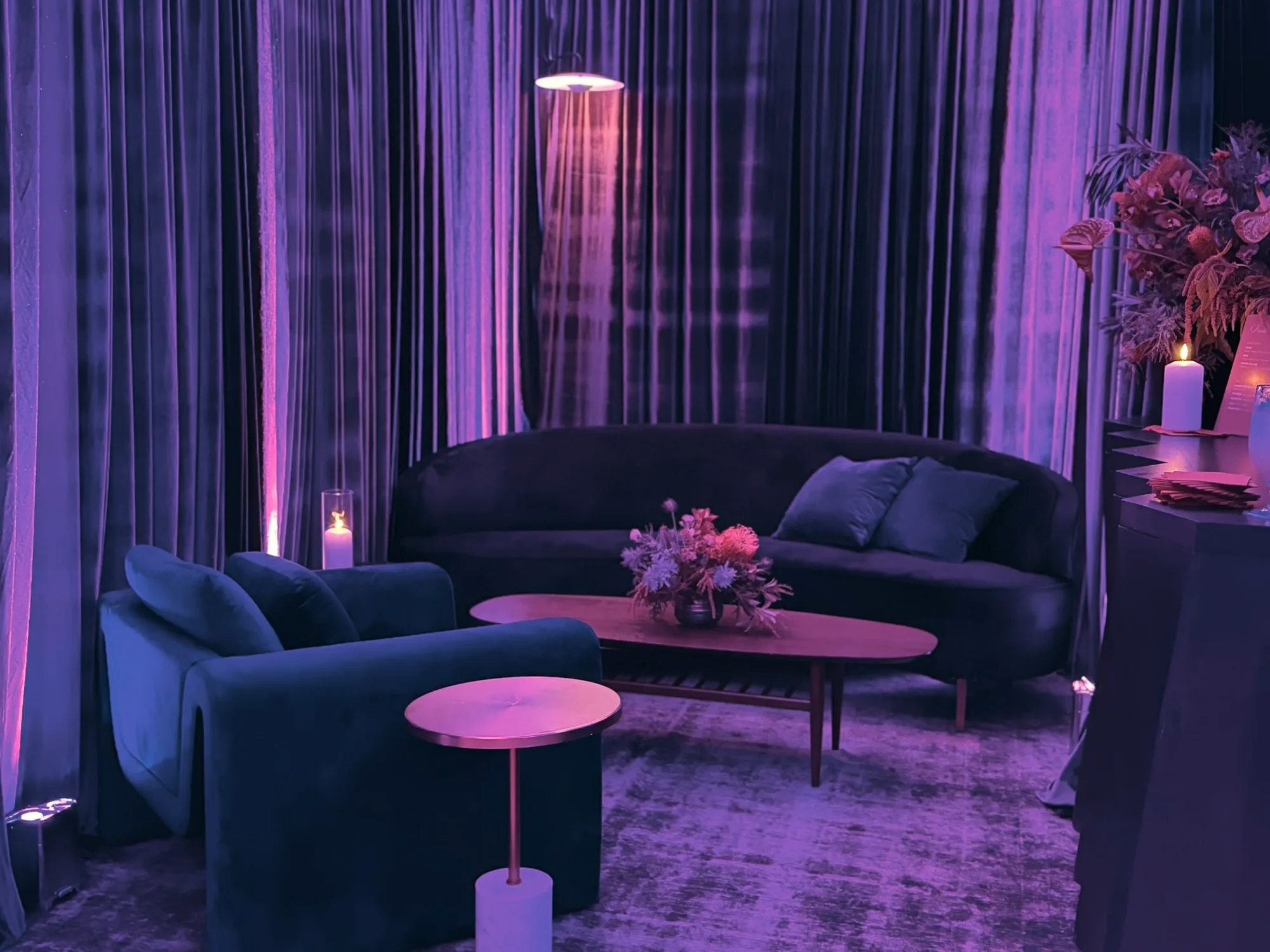 A curved sofa, armchair and low table among tall violet curtains, with candles and flowers.
