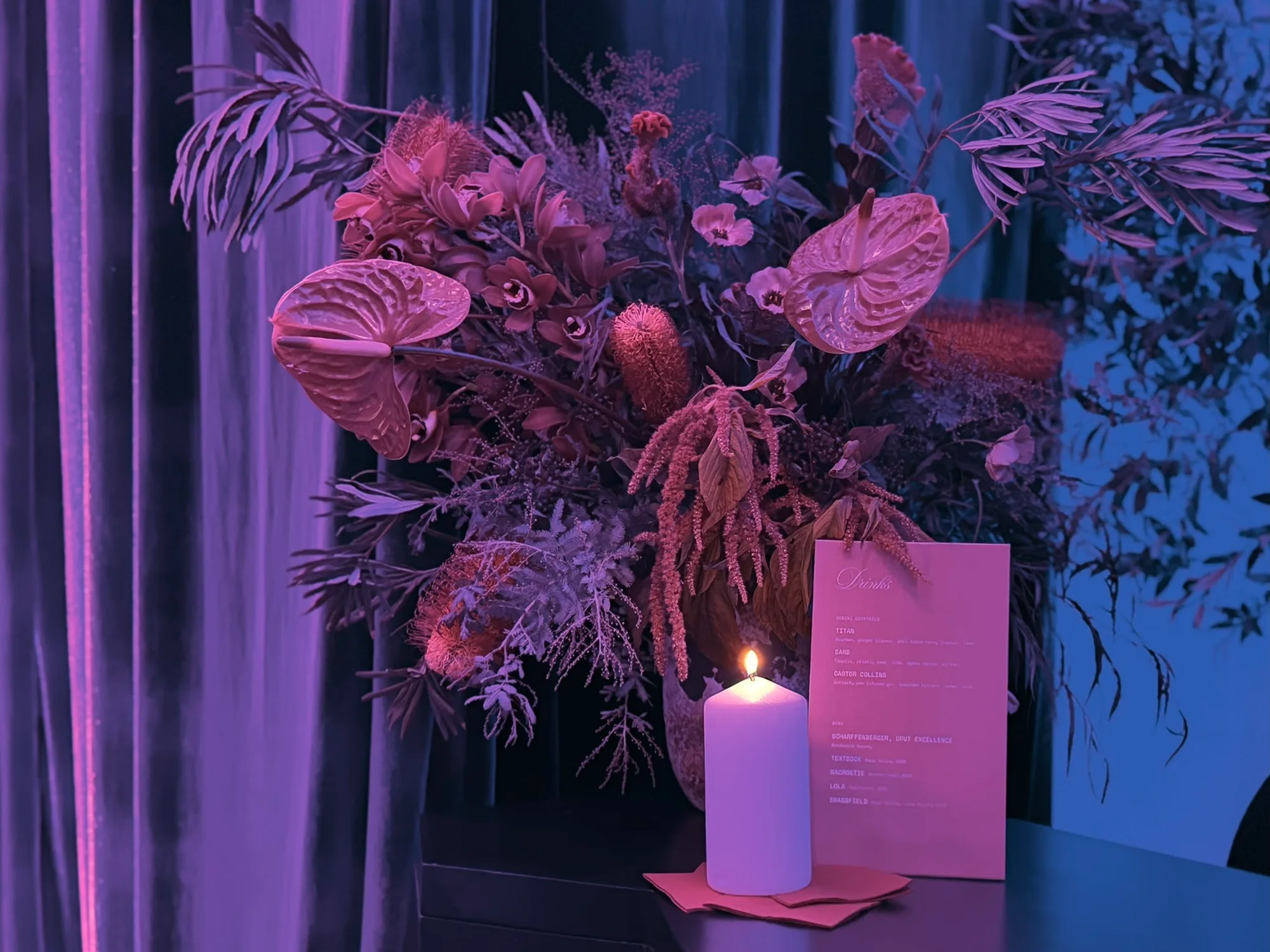Tall floral arrangement with a pillar candle and a pink printed card against violet curtains.
