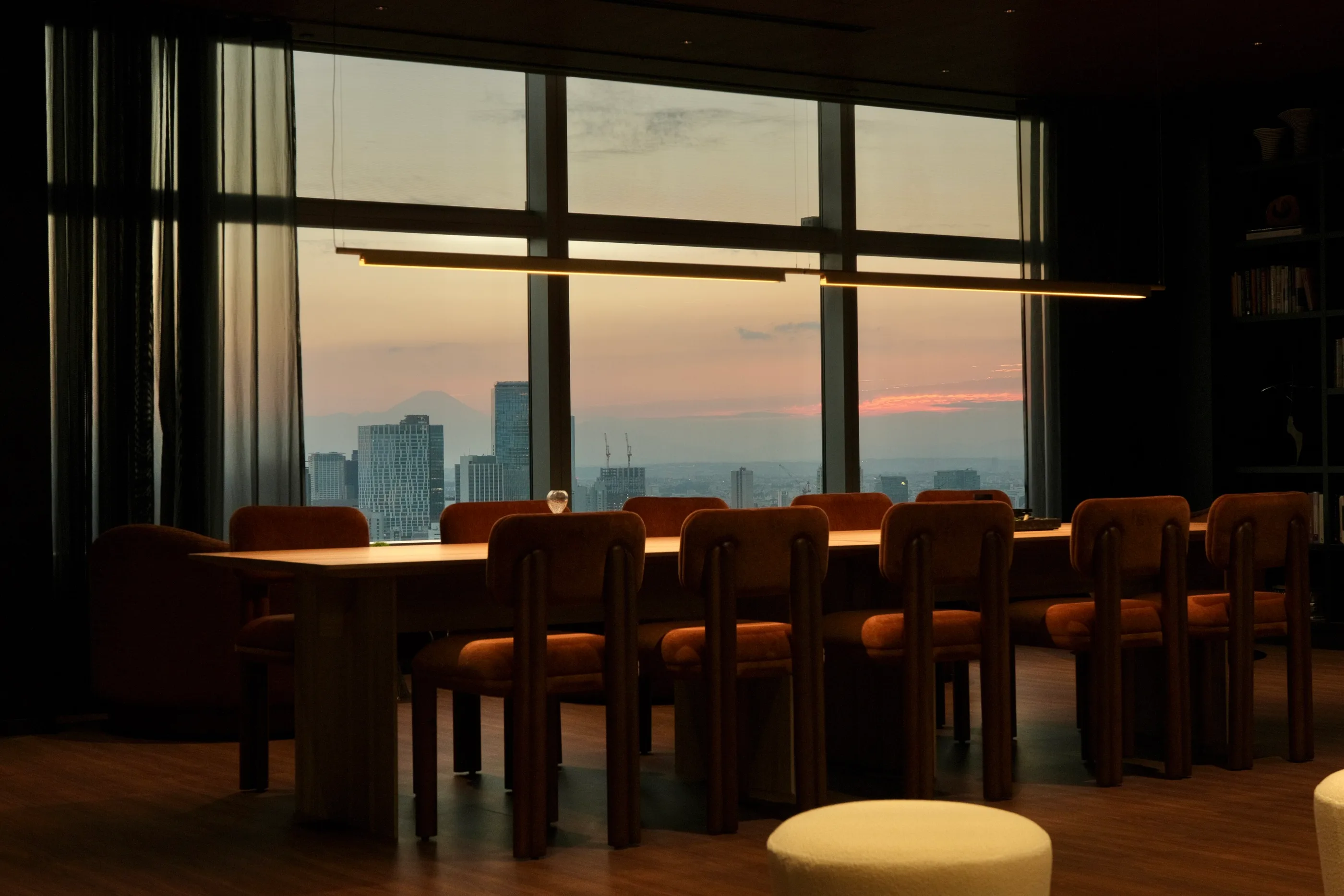 A long table with chairs beside floor-to-ceiling windows at dusk.