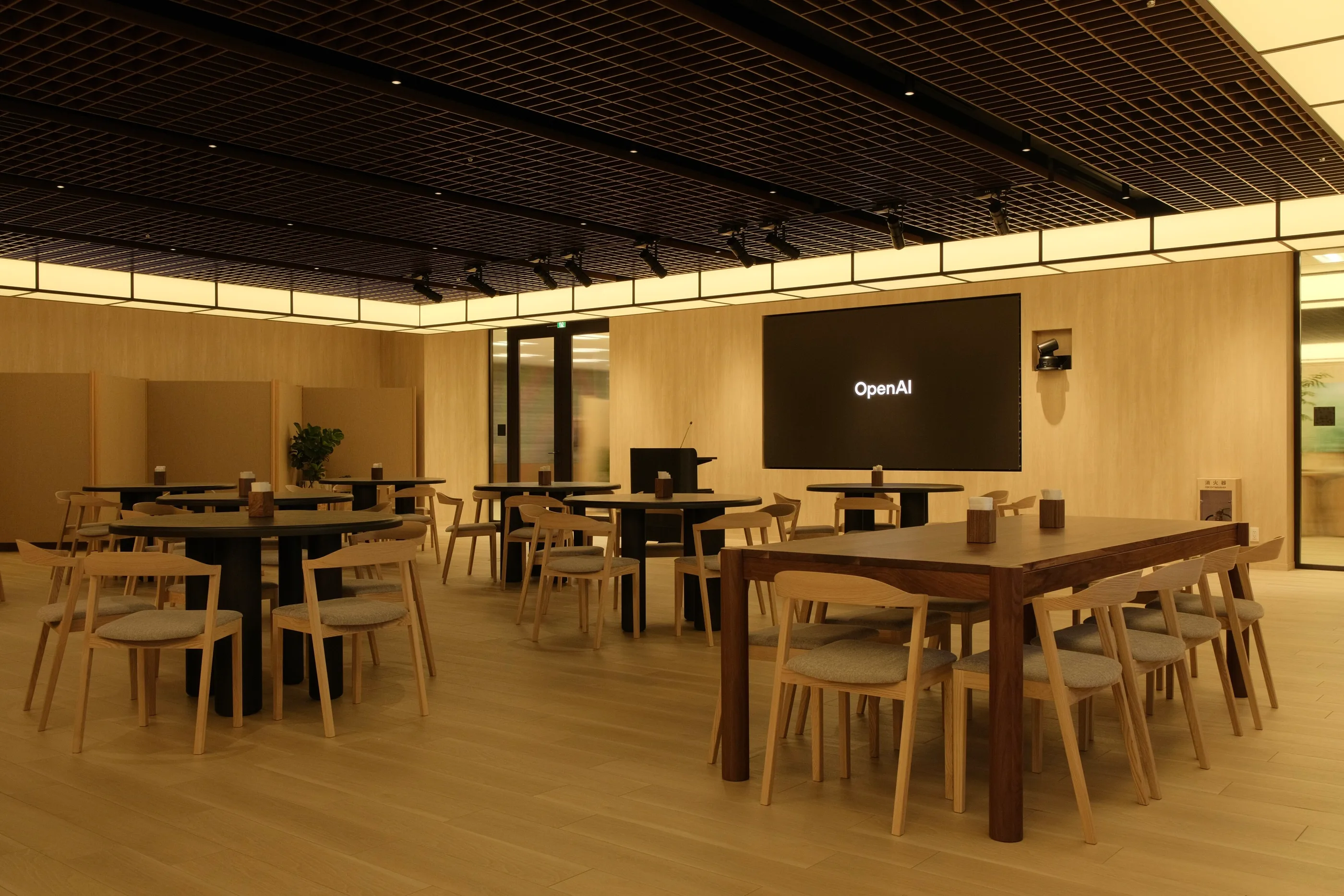 Timber tables and chairs beneath a slatted ceiling with an OpenAI screen on the far wall.