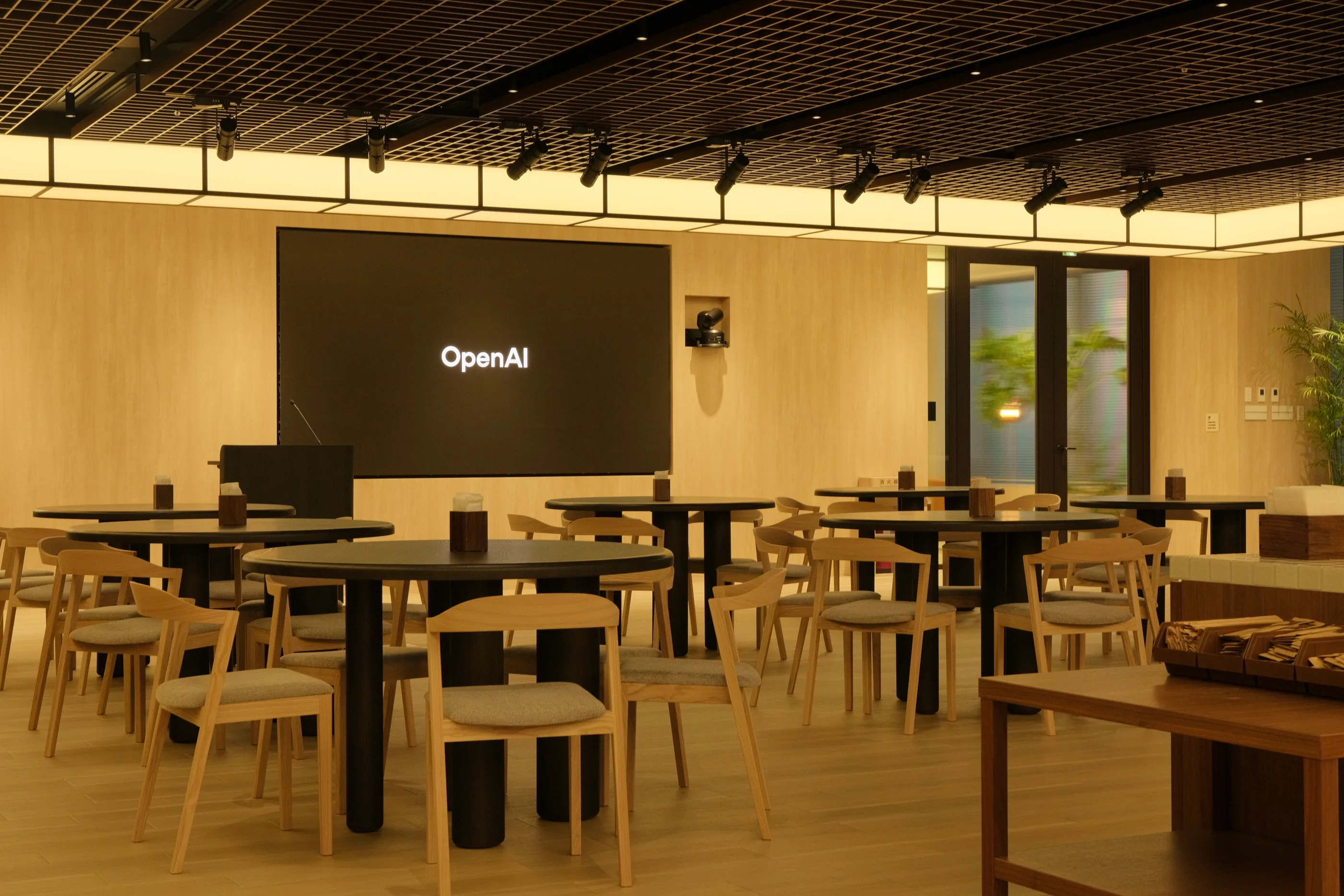 Tables and chairs beneath a glowing ceiling edge with a screen and plants.