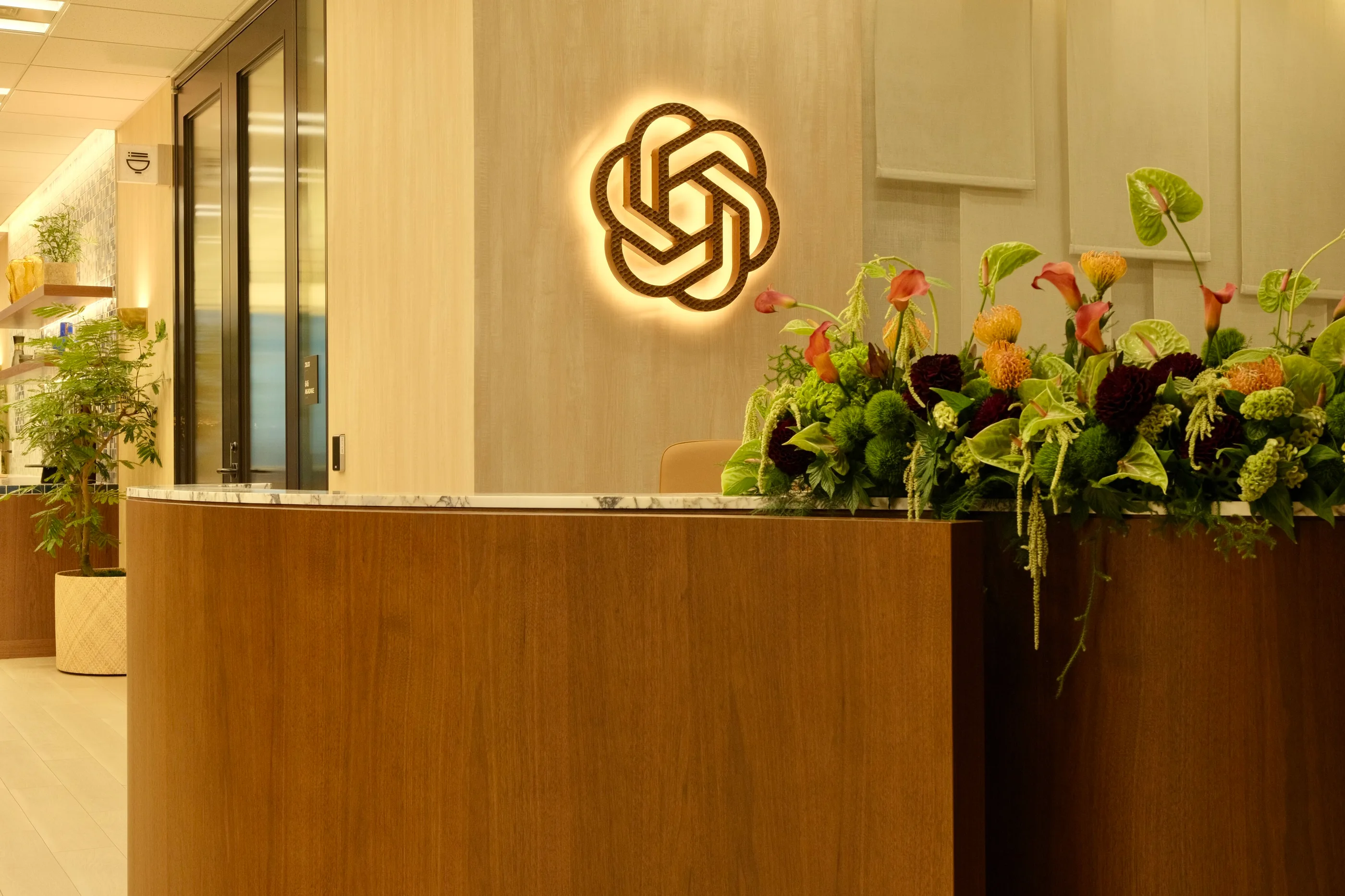 Tokyo reception desk in timber with the OpenAI mark glowing on the wall behind a floral arrangement.