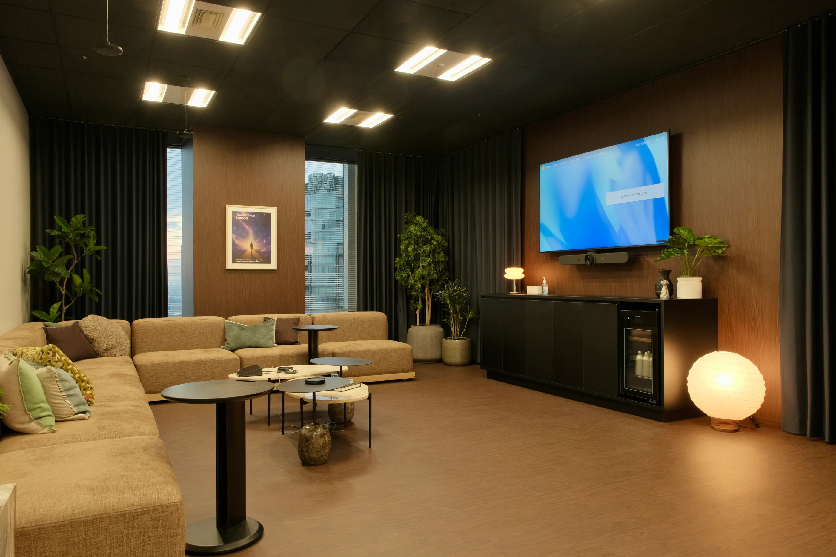 A lounge with a long sofa, a lit floor lamp and a wall display in a dark-walled room.