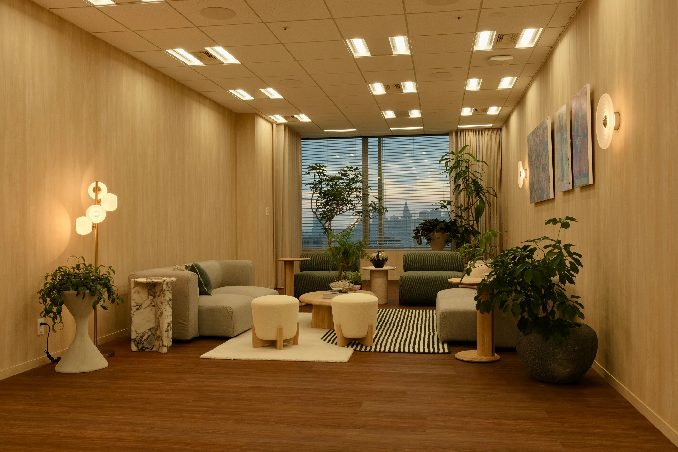 Soft seating, plants and a round table beside tall city-view windows.