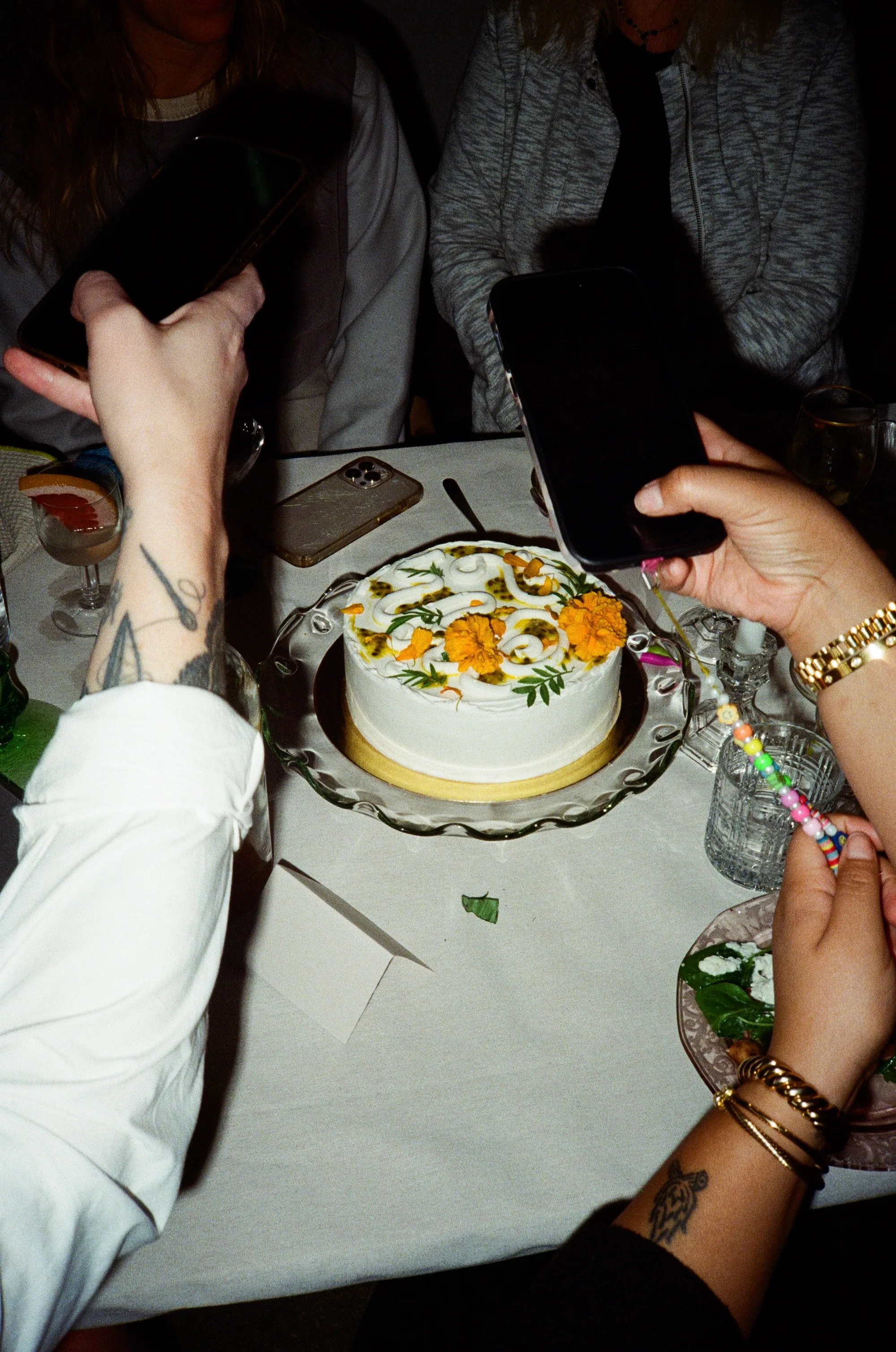 A white cake decorated with orange flowers on a table, with several hands around it.
