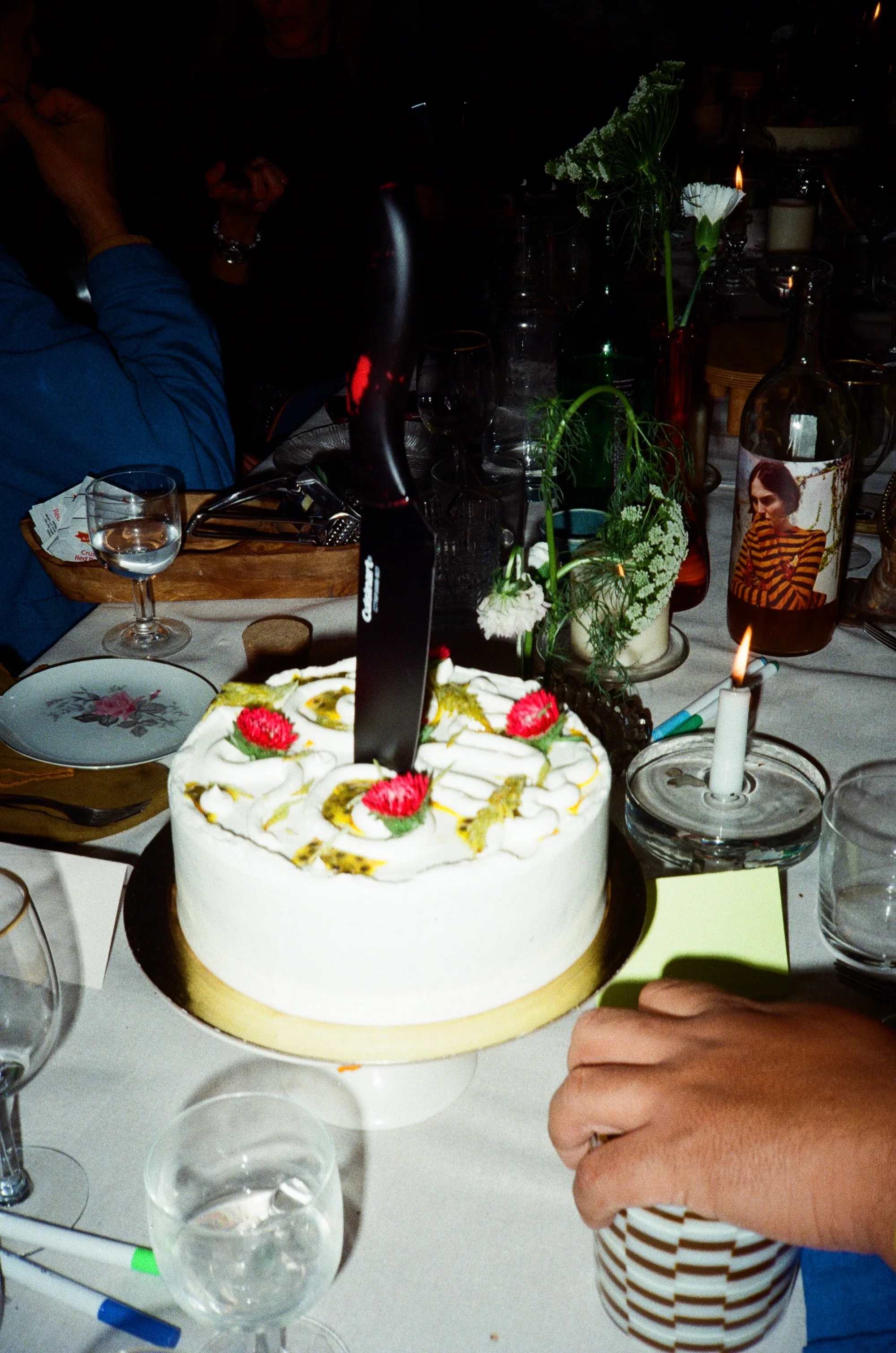 A white cake with strawberries and a candle, a knife held above it, among glasses and bottles.