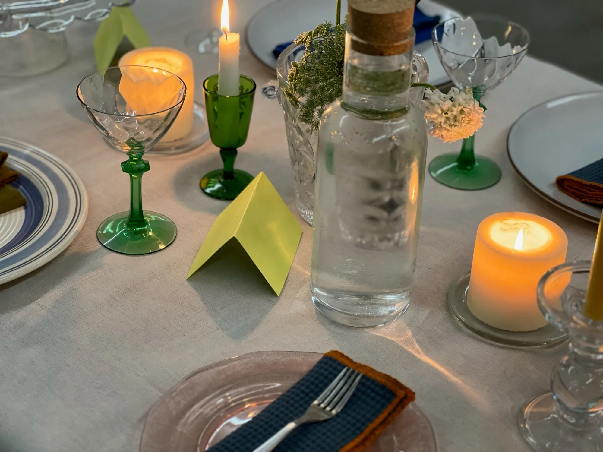Close view of candles in glass holders, green stemware, a clear bottle, and a folded place card.