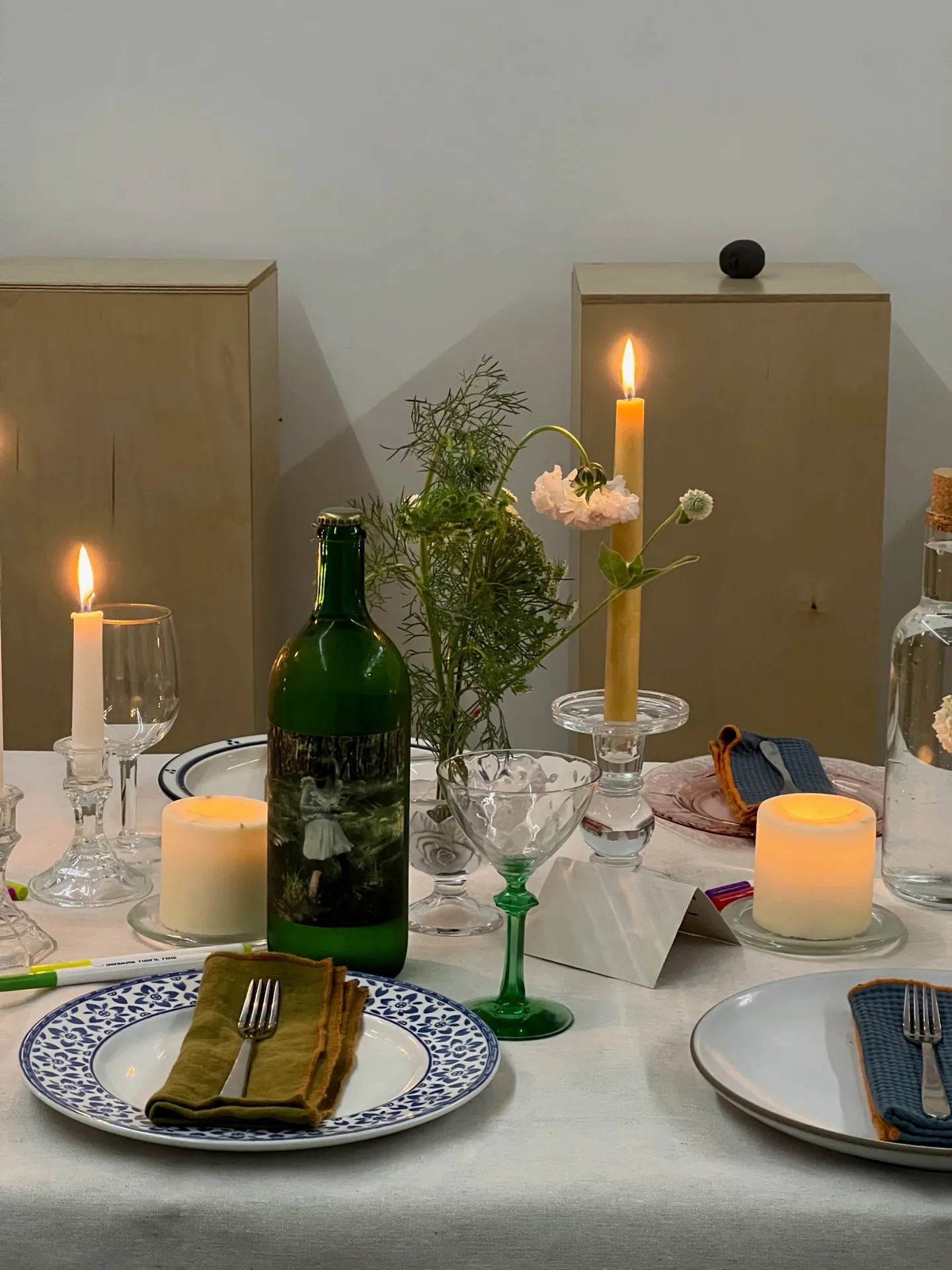 A table with a tall candle, a green wine bottle, a small flower vase, and a blue-patterned plate.