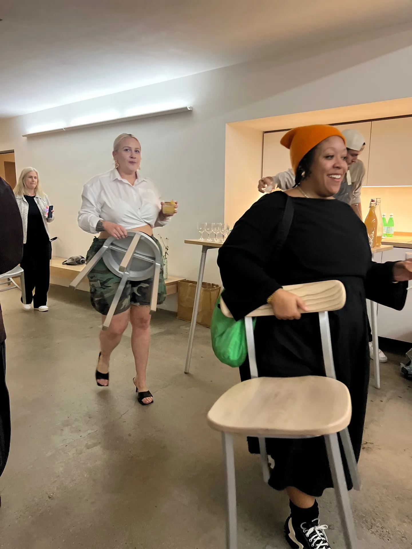 Two people crossing a bright room, one carrying a pale wooden chair.