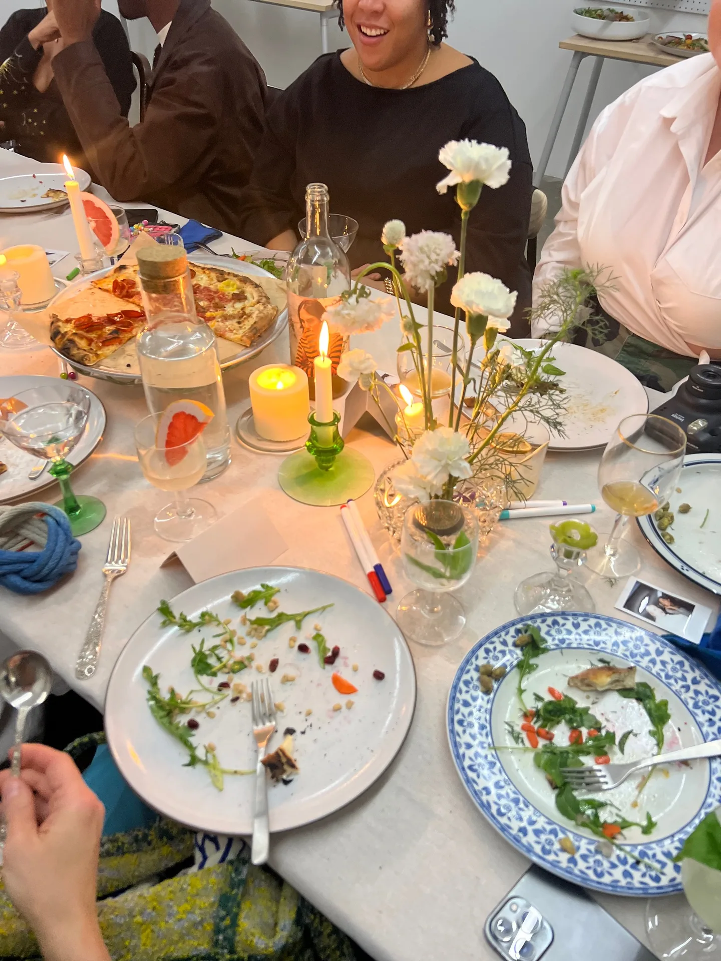 Used plates with greens, candles, and white carnations on a table, and guests laughing in the background.