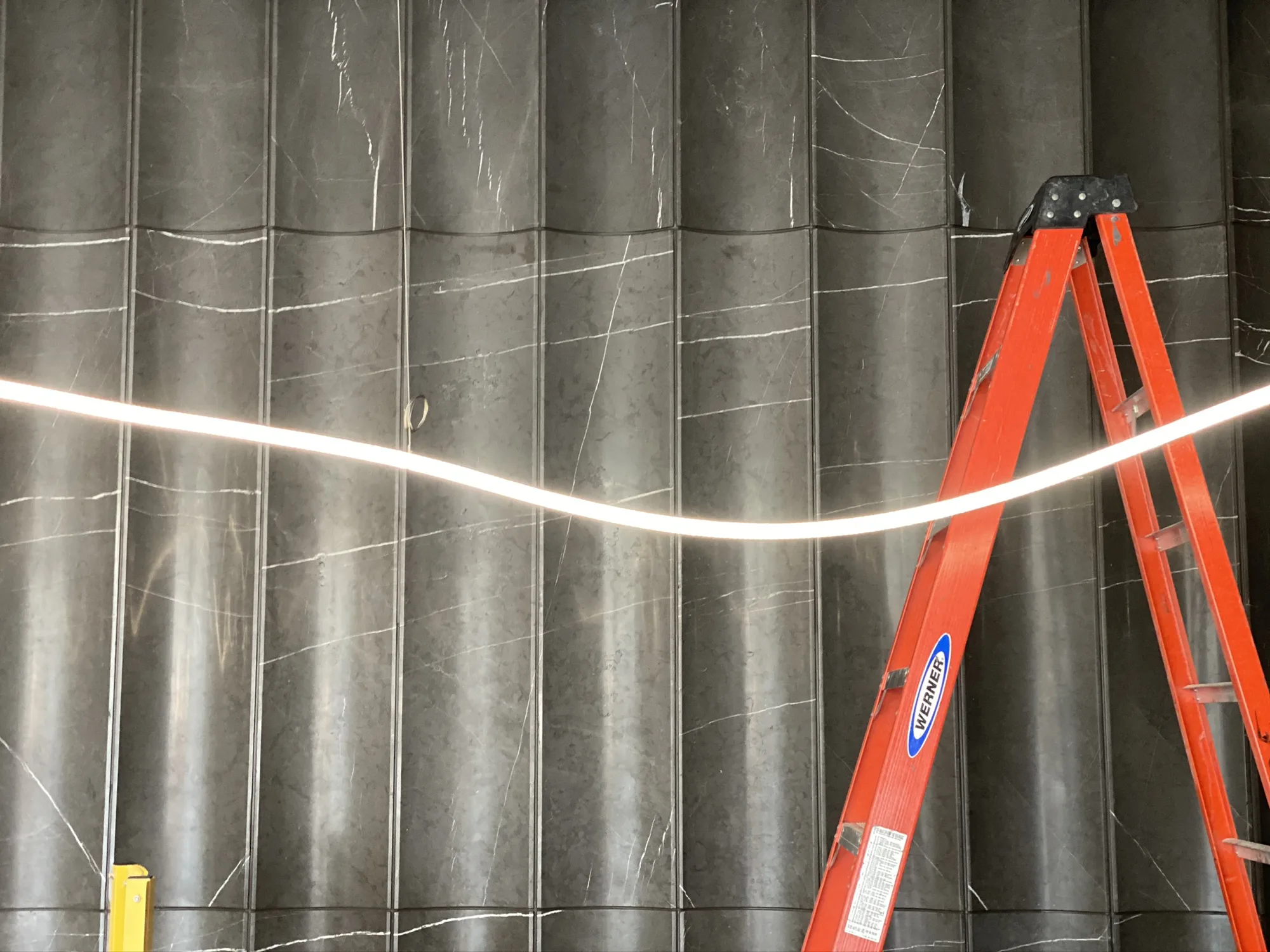 A white line of light curving across dark veined stone panels, with an orange ladder at the right.