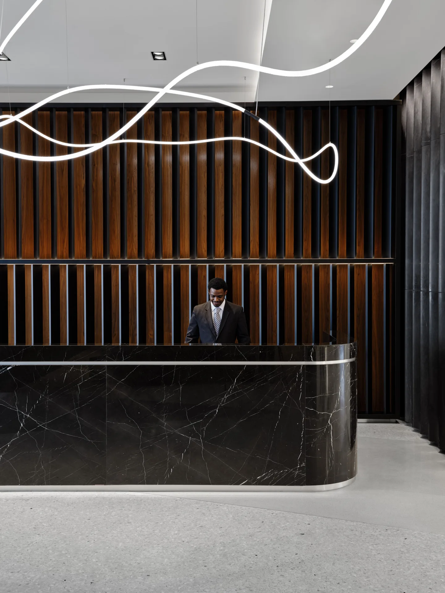 A reception desk of black stone with a person behind it, against vertical timber slats and looping white light lines overhead.