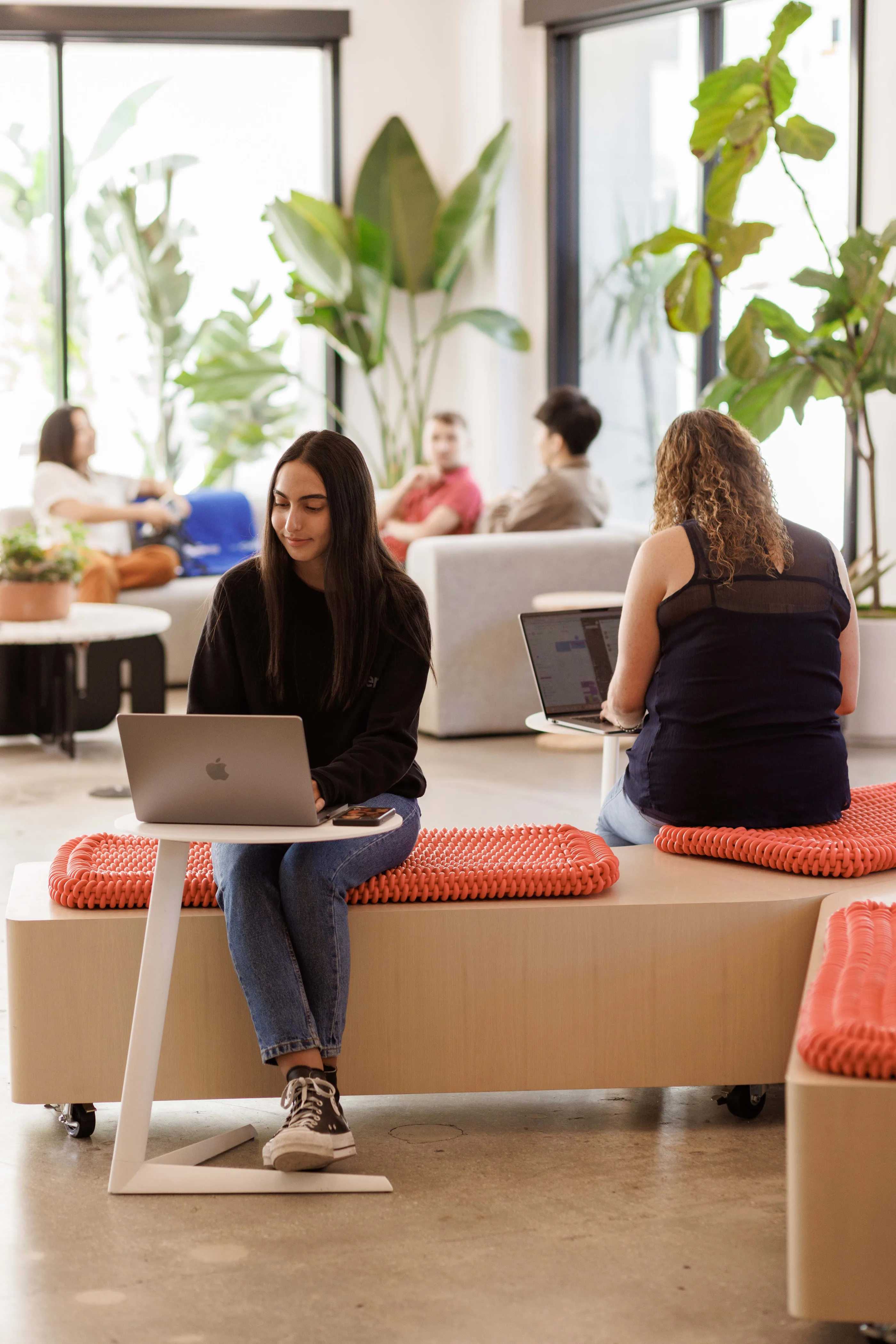 Bright office with people on a pale bench with orange cushions, a laptop on a rolling table, and large plants.