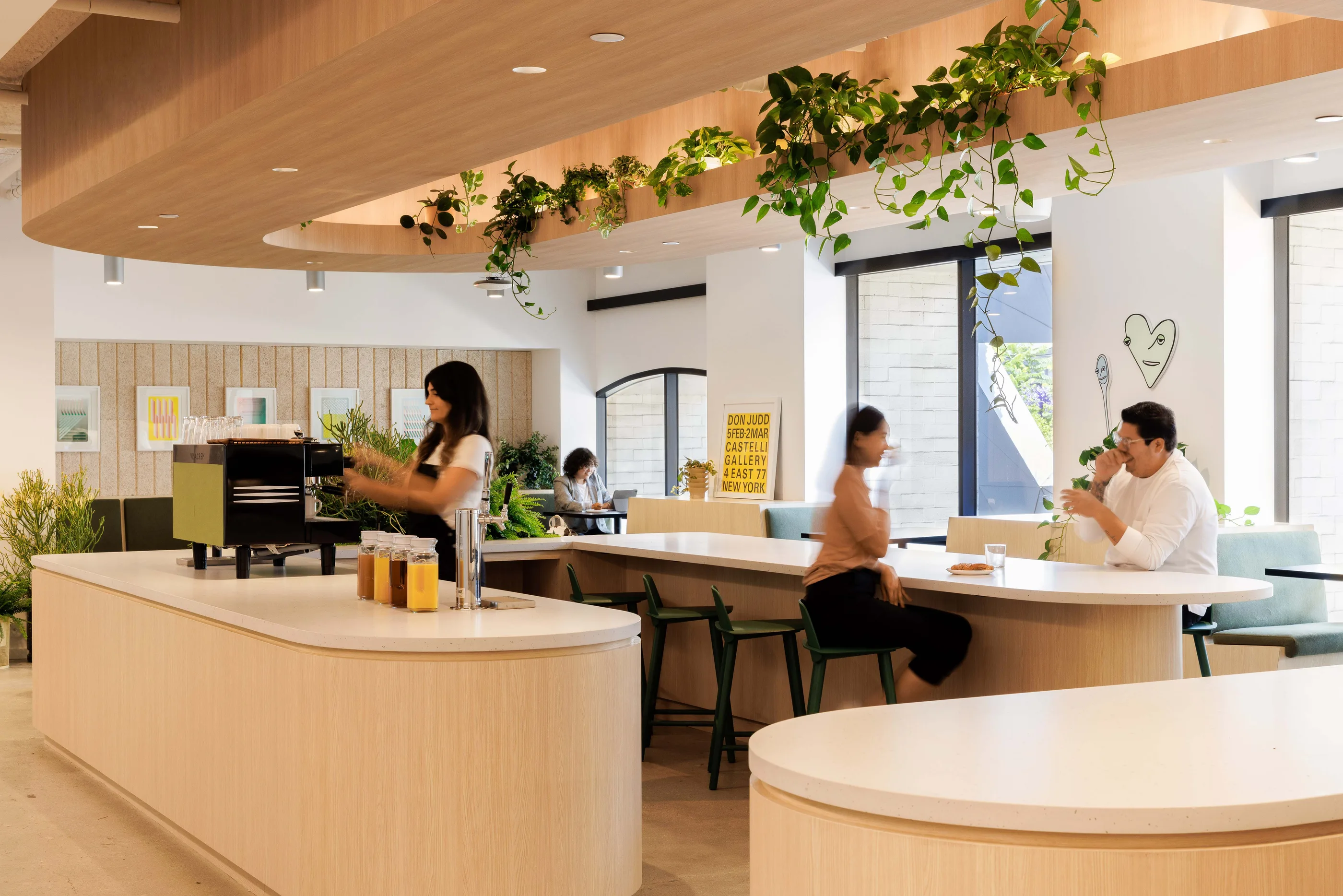 Cafe space with a curved counter, trailing plants above, and people at tables near large windows.