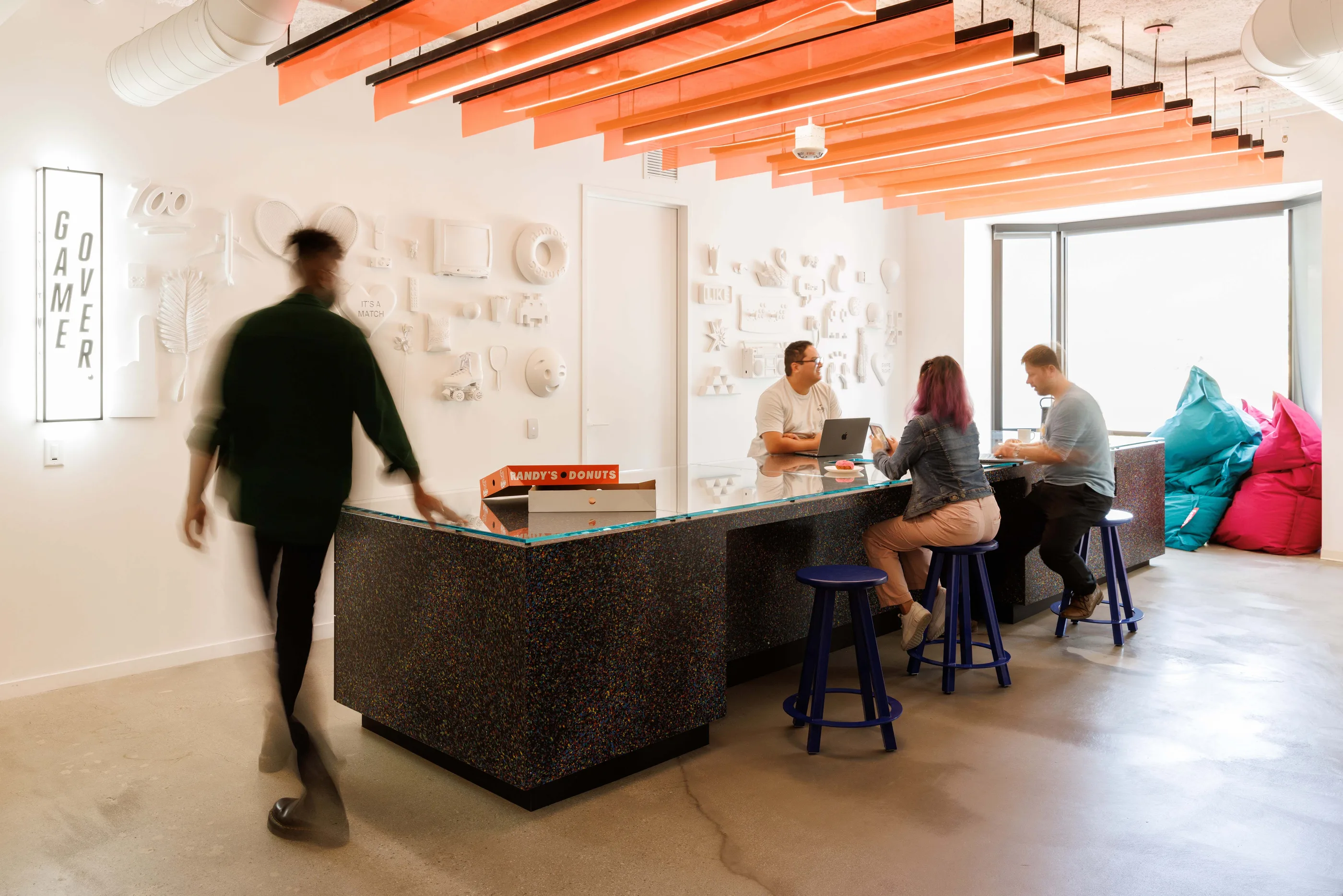 Room with an orange slatted ceiling, a terrazzo counter with stools, beanbags, and a white relief wall.