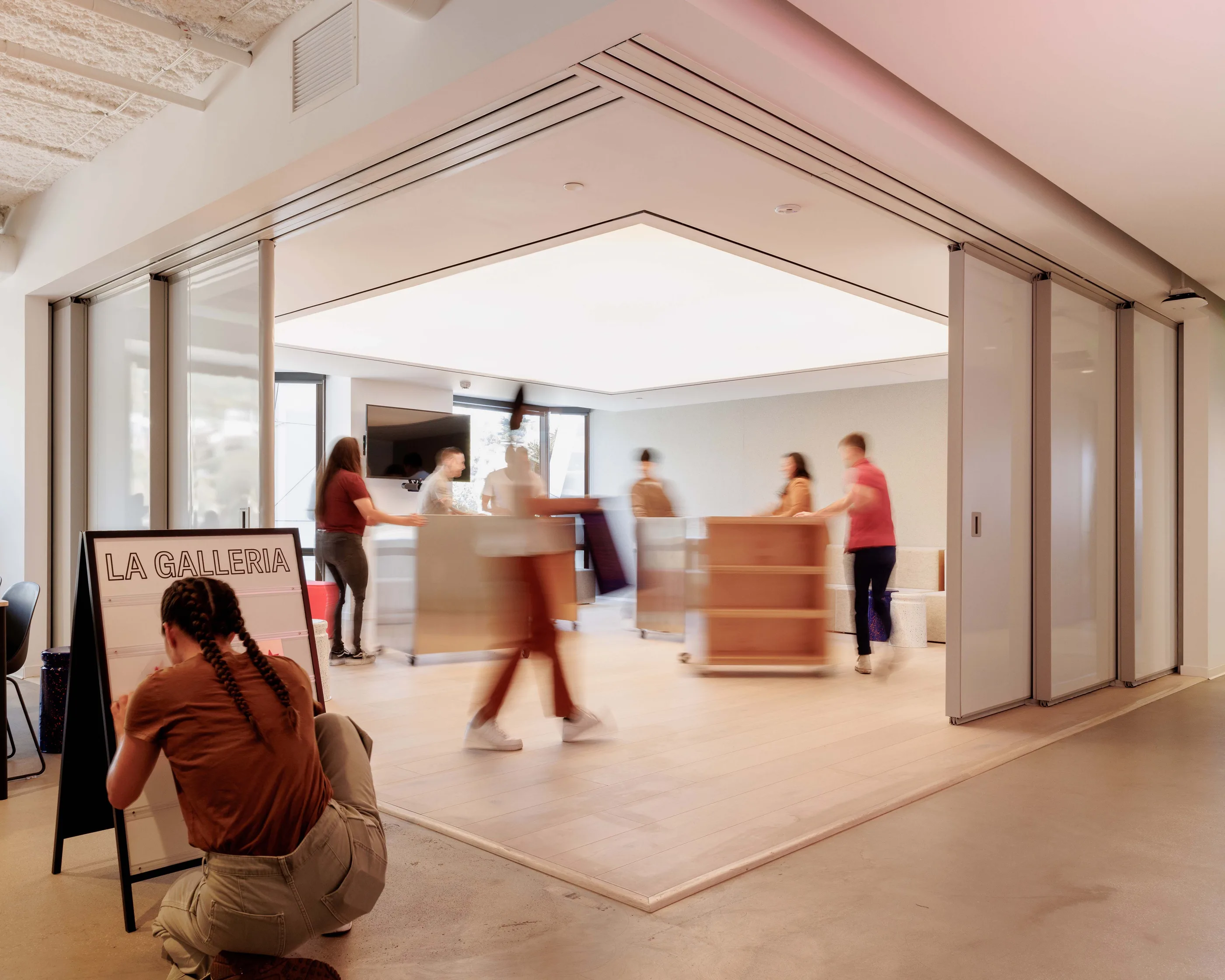 View through an open sliding wall into a gallery space with an easel sign reading La Galleria and people moving inside.
